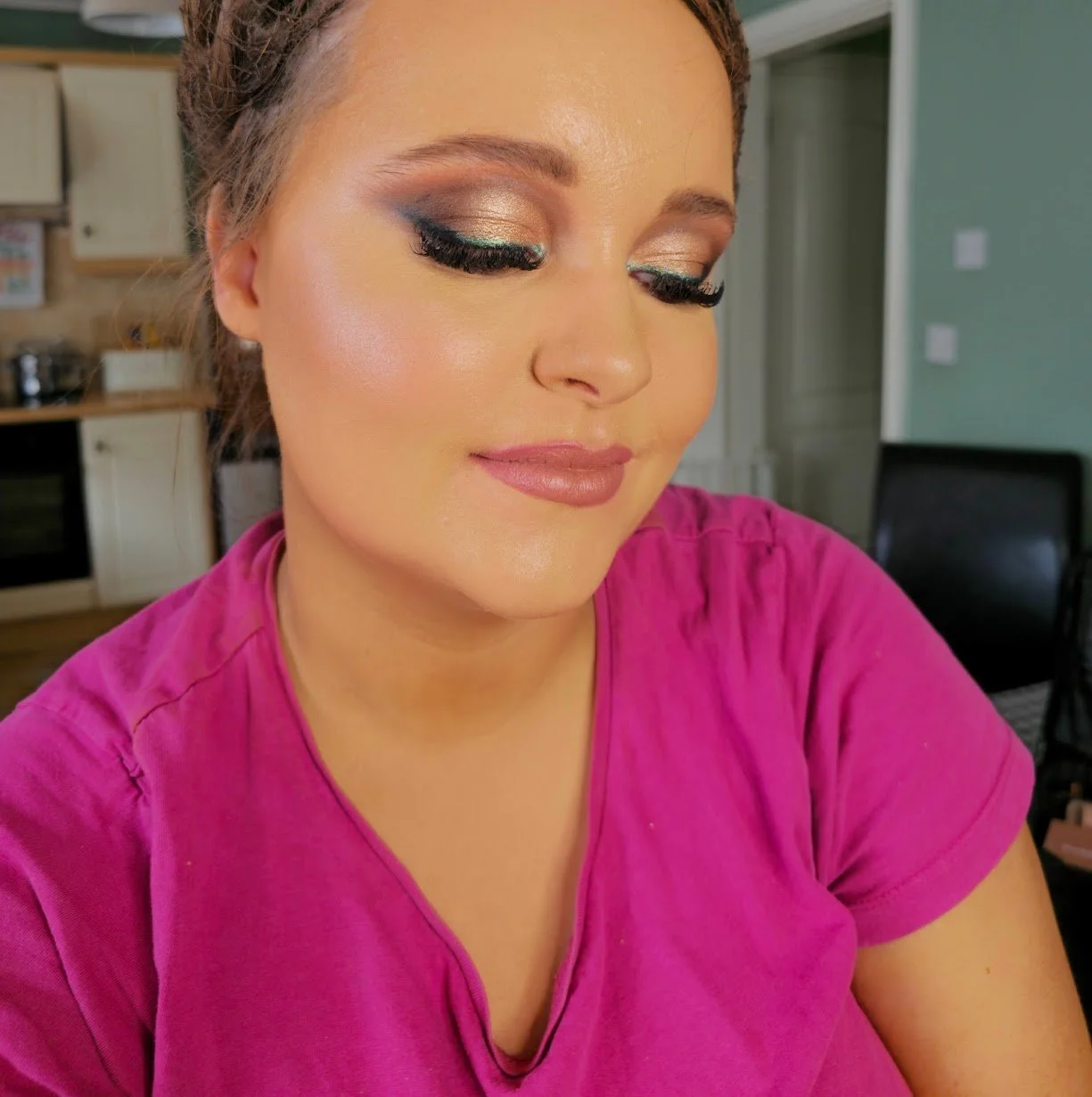 A woman with makeup including gold eyeshadow, thick black eyeliner, and pink lipstick, wearing a pink shirt, with a kitchen in the background.