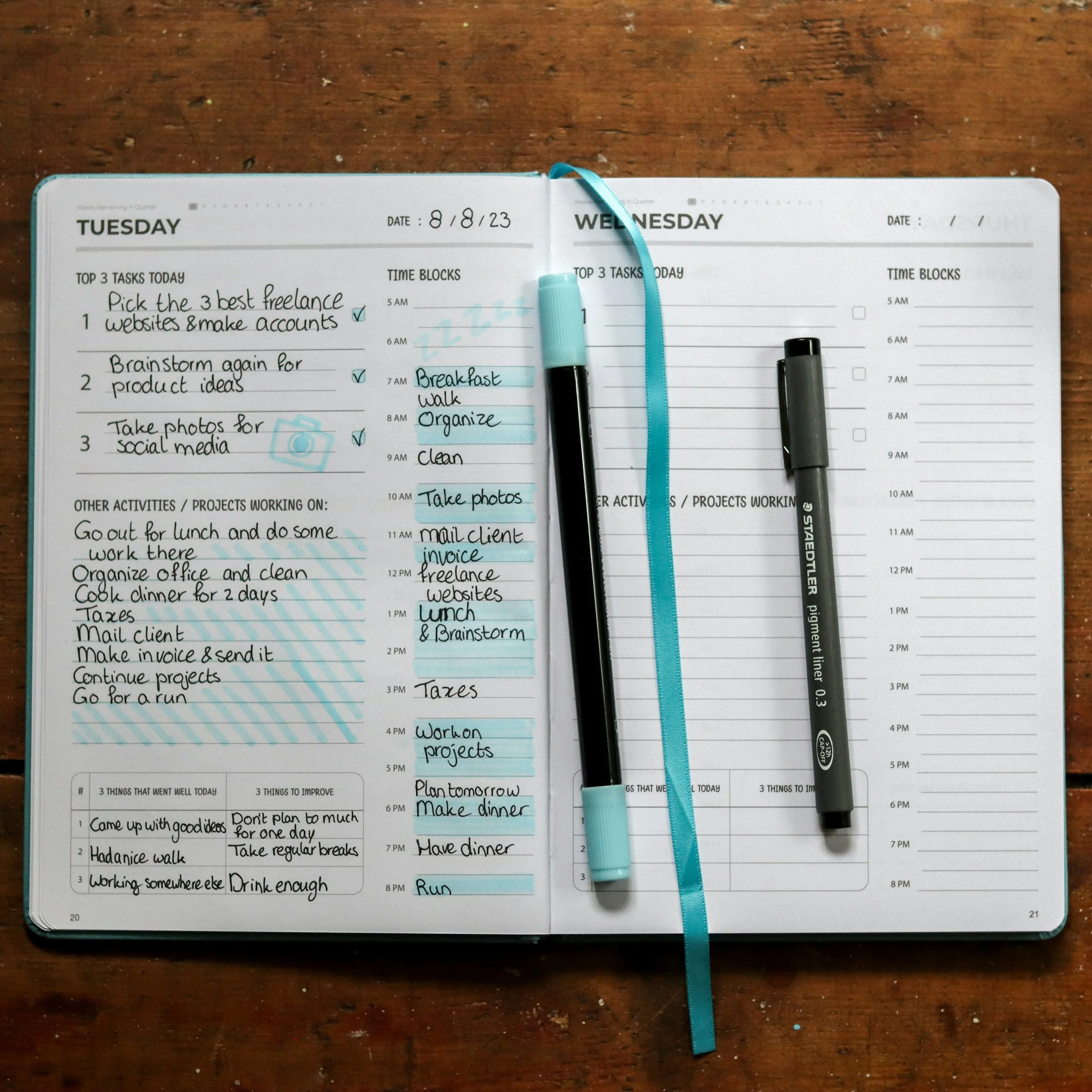 Open planner with handwritten tasks for Tuesday and Wednesday, two pens placed on the pages, and a blue ribbon bookmark.