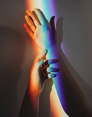 Two hands with rainbow-colored shadows on a plain wall, one hand touching the wrist of the other.