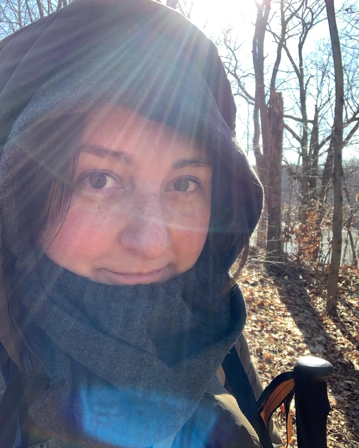 A woman bundled up in winter clothing, including a hood and scarf, taking a selfie outdoors in a wooded area on a sunny day with sunlight streaming through the trees.