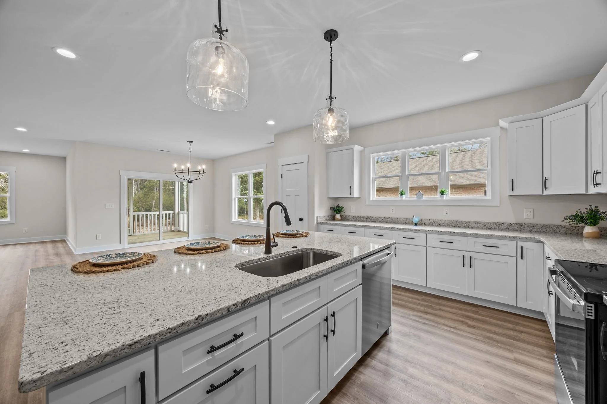 Modern, empty kitchen with white cabinets, granite countertops, stainless steel sink and appliances, wood flooring, and large windows, open to a dining and living space with pendant lights and sliding door to outdoor deck.