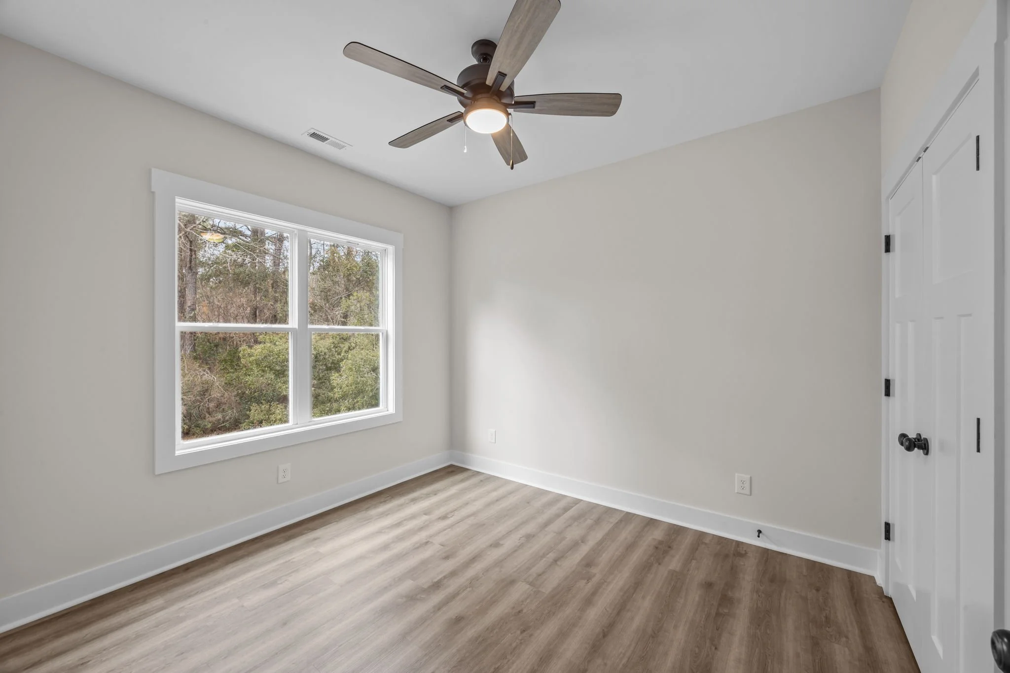 Empty room with white walls, a large window, a ceiling fan, wood flooring, and a closed closet door.
