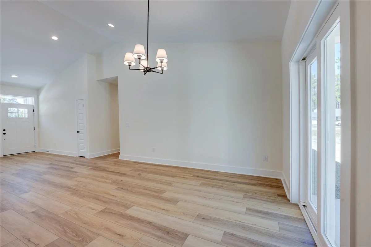 Empty living room with white walls, light wood flooring, a chandelier, and sliding glass doors leading outside.