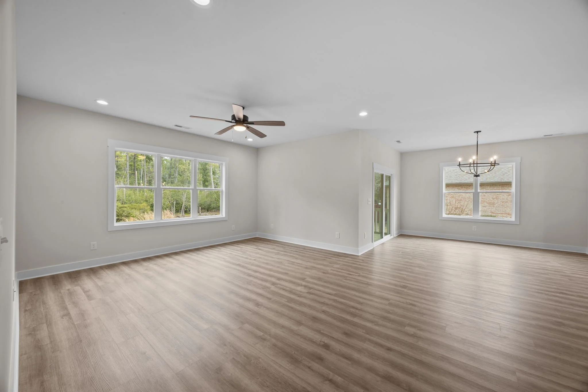 Empty living room with large windows, ceiling fan, light-colored walls, and light wood flooring.