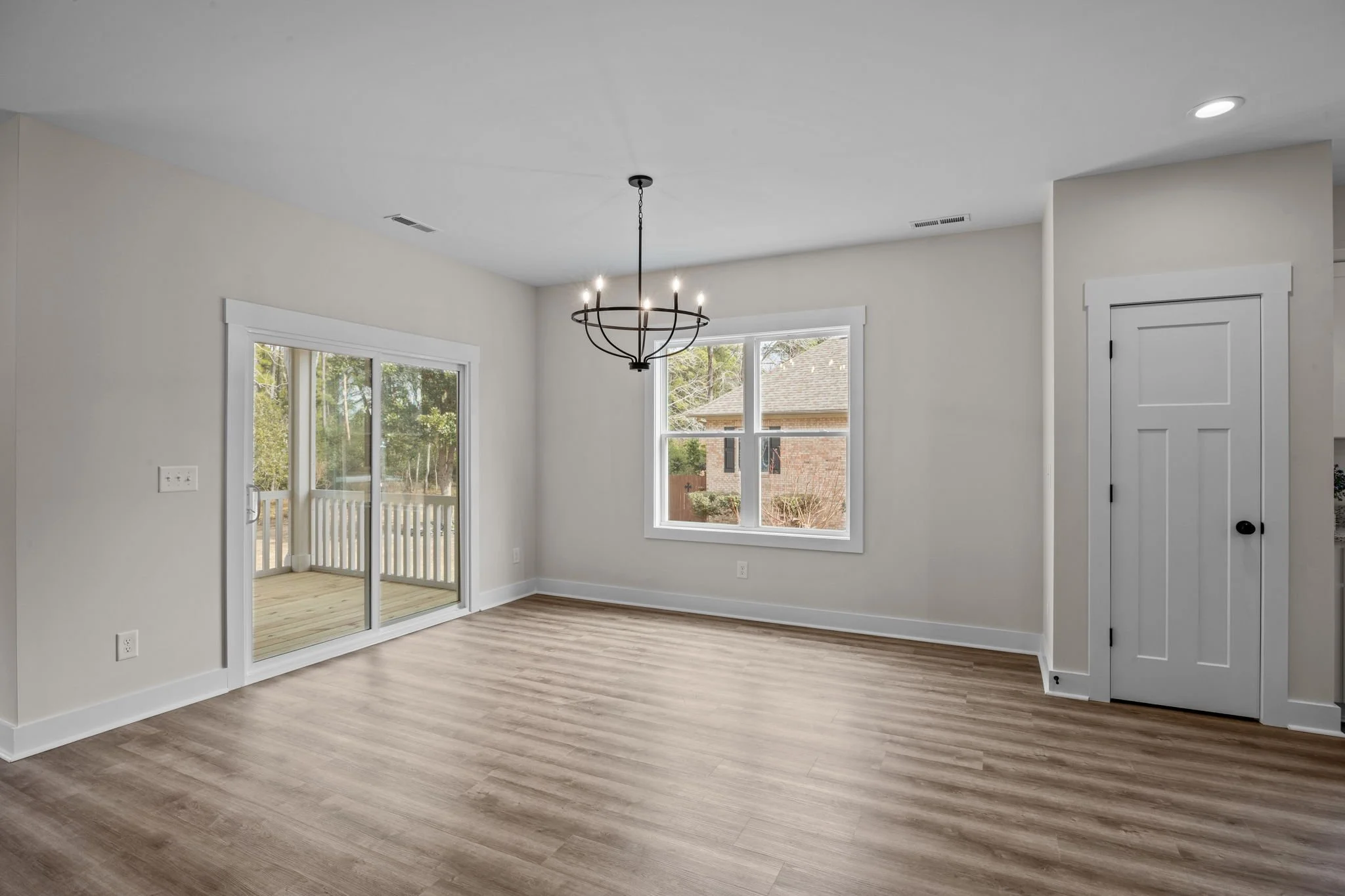 Empty living room with wood flooring, white walls, a chandelier, a sliding glass door leading to a balcony, and a window showing trees and neighboring houses.