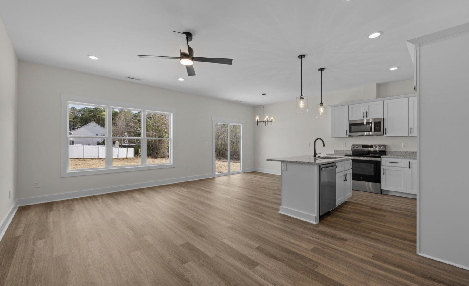 Empty living and kitchen area in a new home with large windows, hardwood floors, white cabinets, black appliances, and modern light fixtures