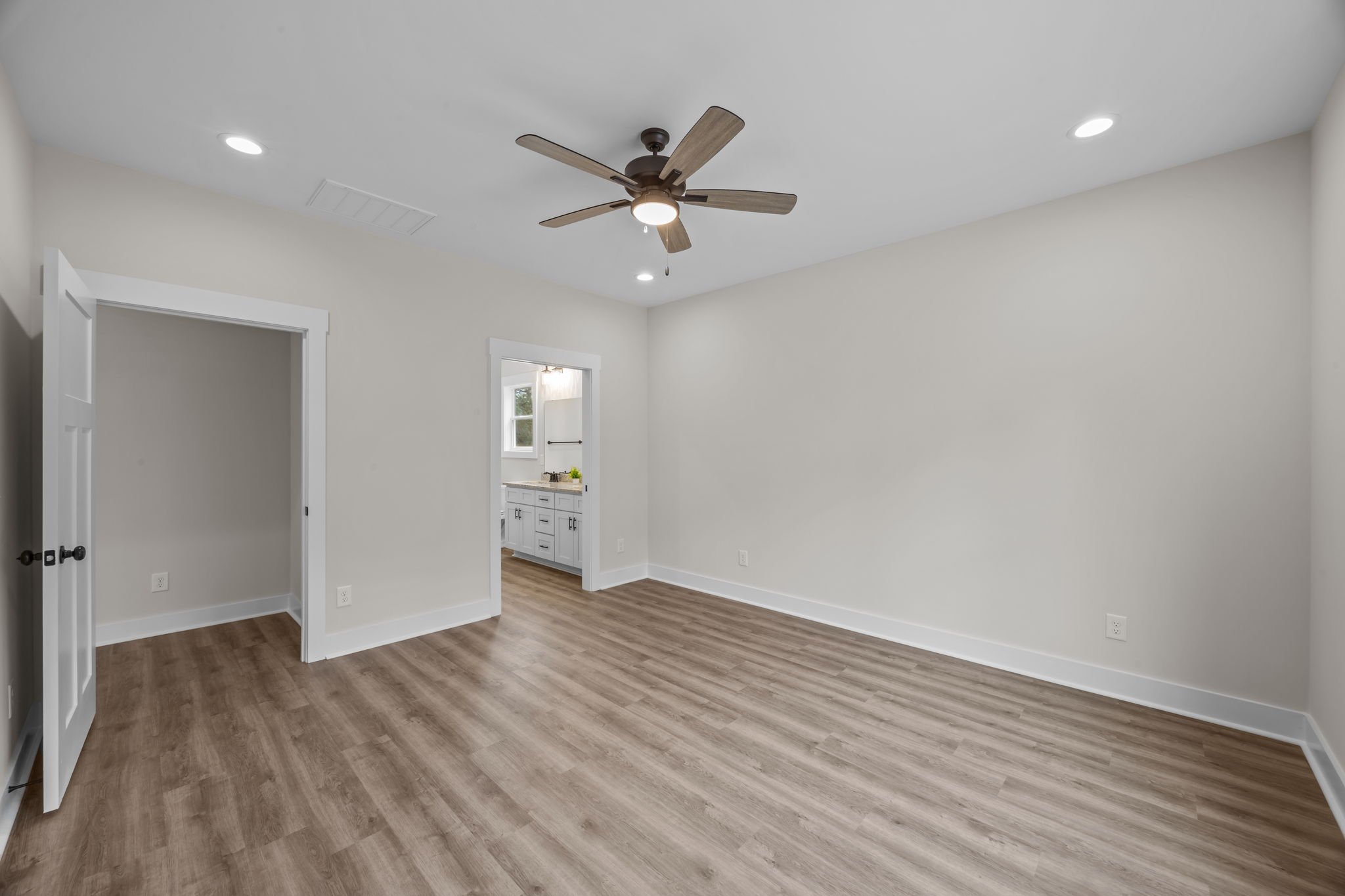 Empty room with light wood flooring, white walls, a ceiling fan, and a doorway leading to a bathroom.