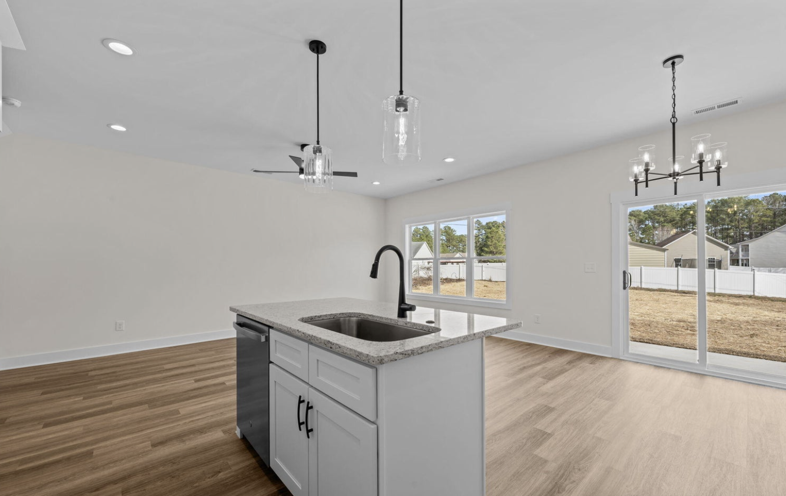 Empty kitchen and dining area with a small island, light wood flooring, white walls, large window, sliding glass door, and modern light fixtures.