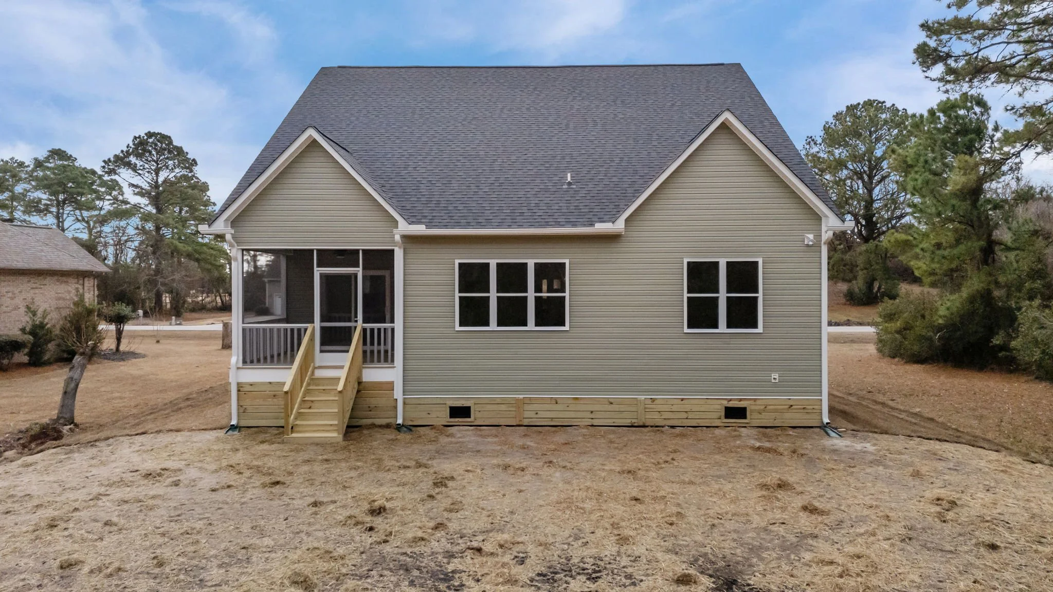 Back view of a house with gray siding, a gabled roof, and a small porch with stairs, surrounded by grass and trees.