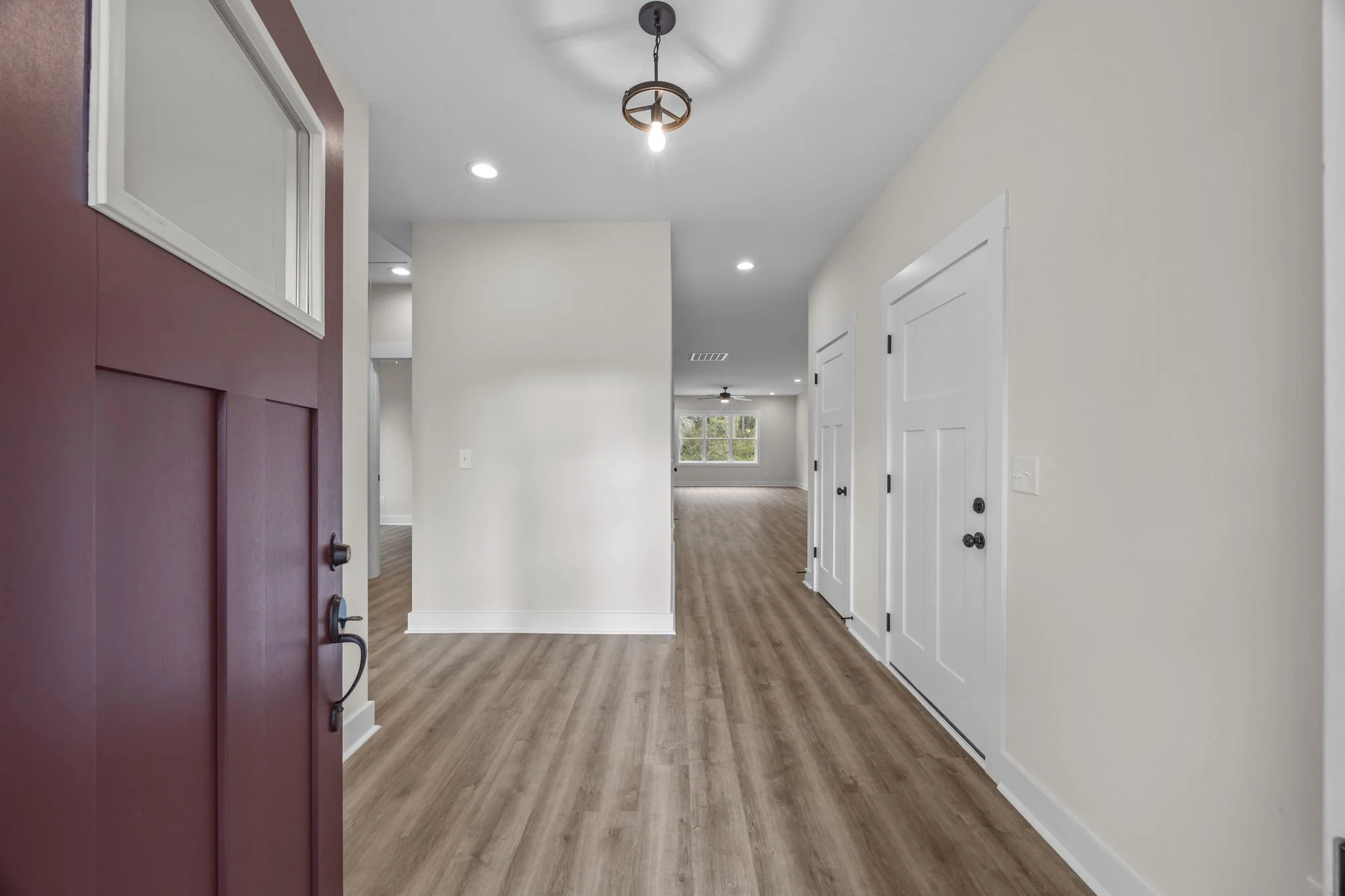 Empty living space with white walls, wooden flooring, white doors, ceiling lights, and a window in the background.
