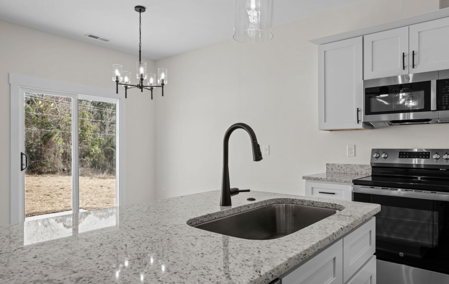 Modern kitchen with white cabinetry, a granite countertop island with a black sink and faucet, stainless steel stove and microwave, and a sliding glass door leading outside. A black chandelier with multiple lights hangs from the ceiling.