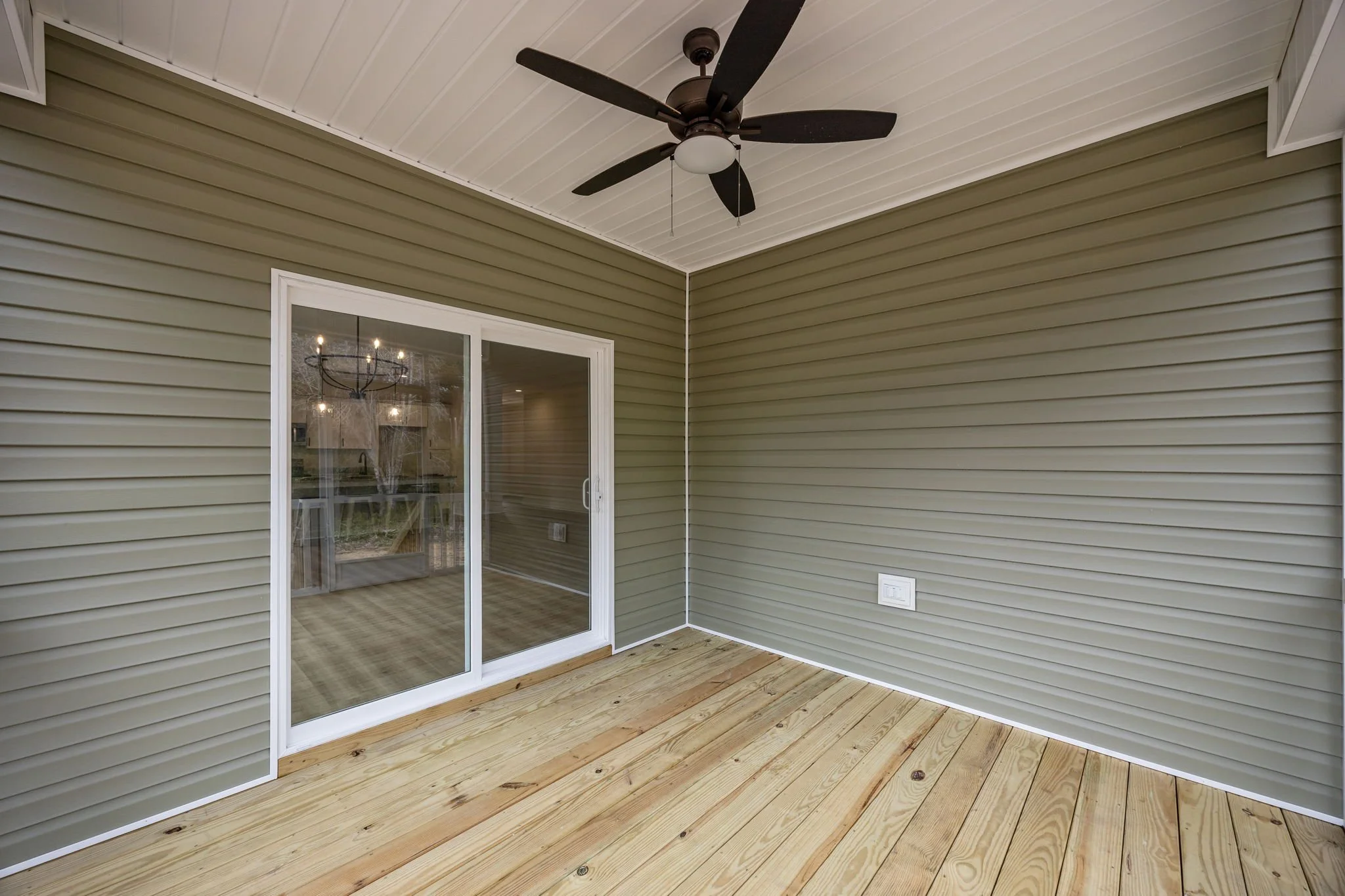 Empty porch area with wooden floor, beige vinyl siding walls, a sliding glass door, a white ceiling, a black ceiling fan, and a chandelier reflection in the glass door.