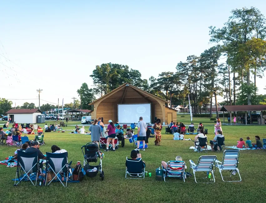 People gathered outdoors on a grassy area watching a movie on a large screen with a wooden pavilion behind it, some sitting in chairs and others on blankets.