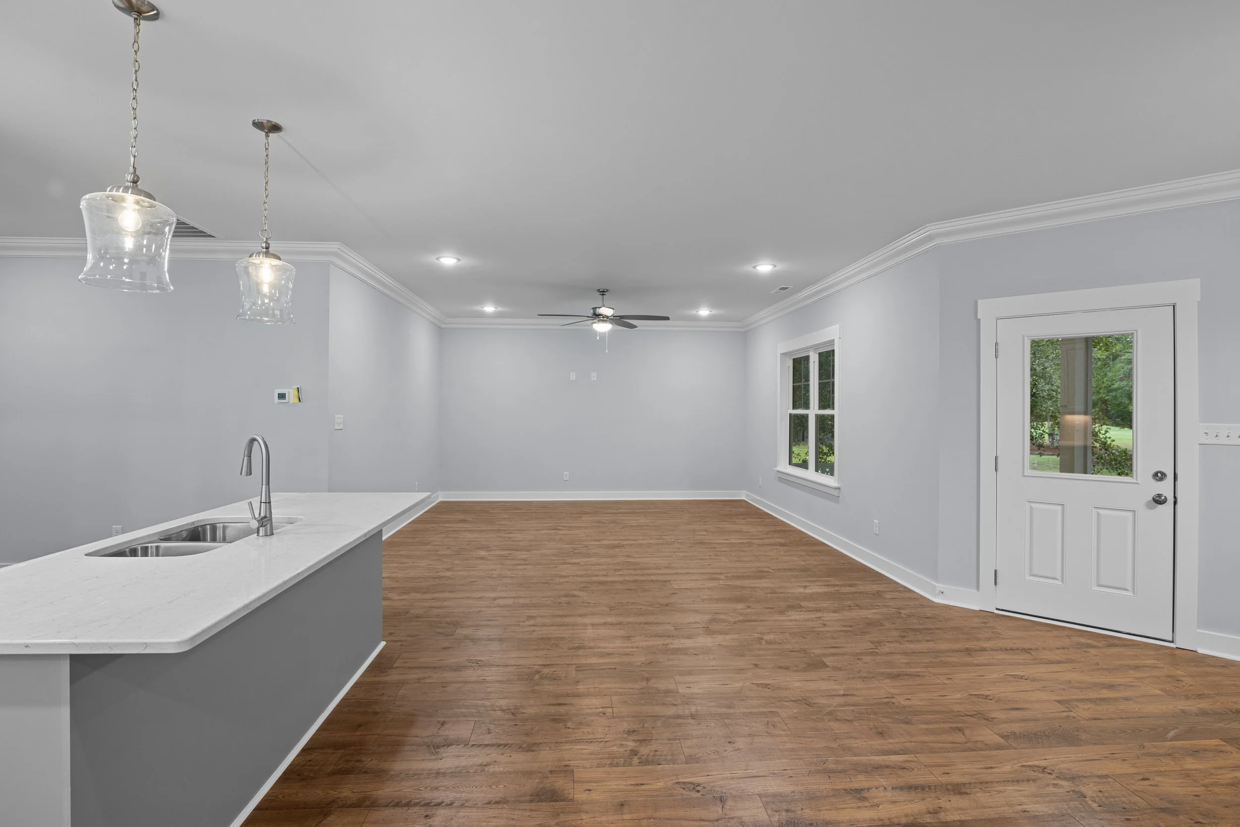 Empty living room with hardwood floors, white walls, a ceiling fan, windows, and a door leading outside.
