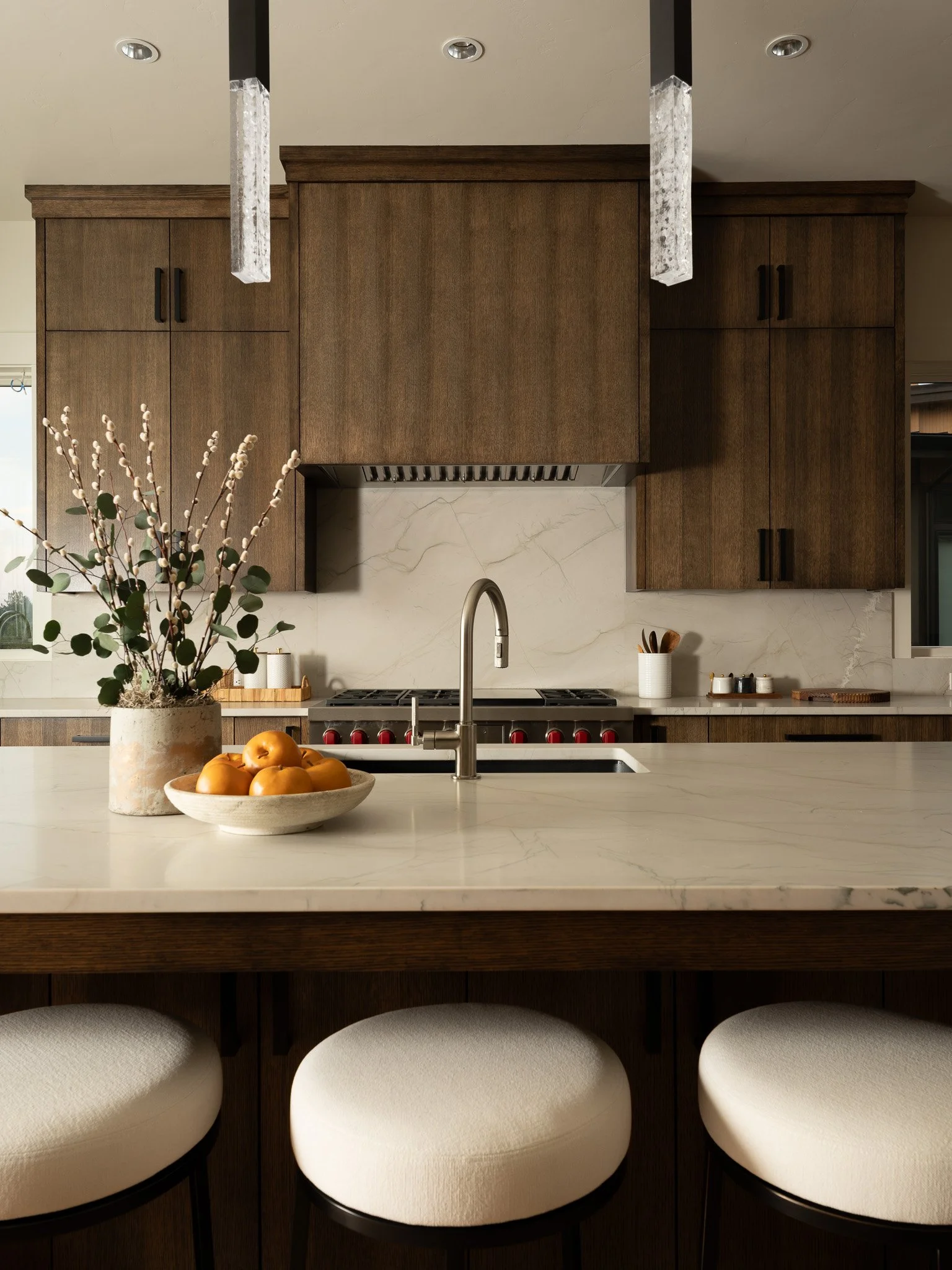 A modern kitchen with wooden cabinets, a marble countertop island, and a white sink with a silver faucet. There are seven beige cushioned bar stools along the island. A chopping board and containers are on the counter, with a bowl of yellow apples an