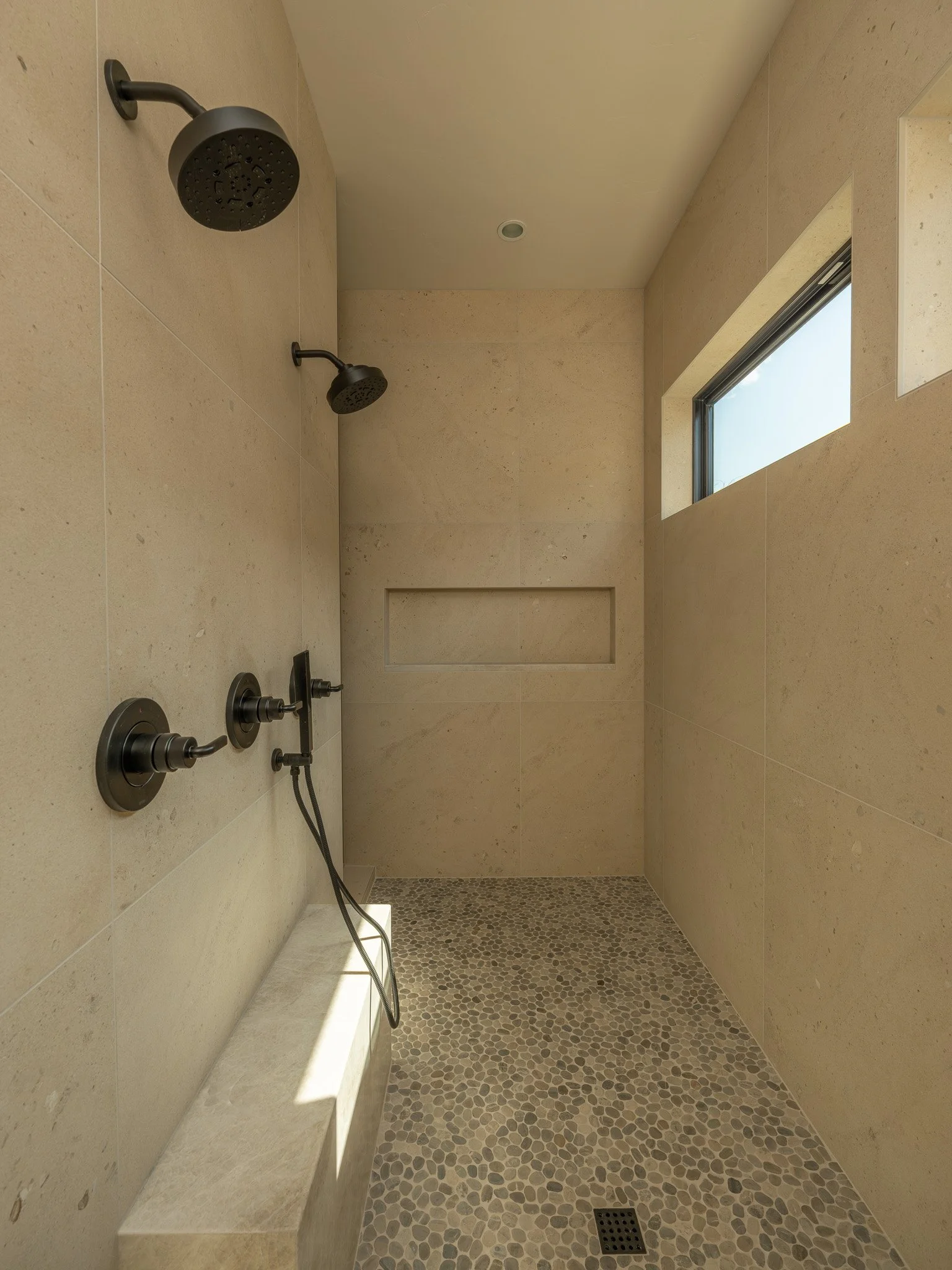 A modern walk-in shower with beige tiled walls, two black showerheads, and a pebble stone floor with a drain.