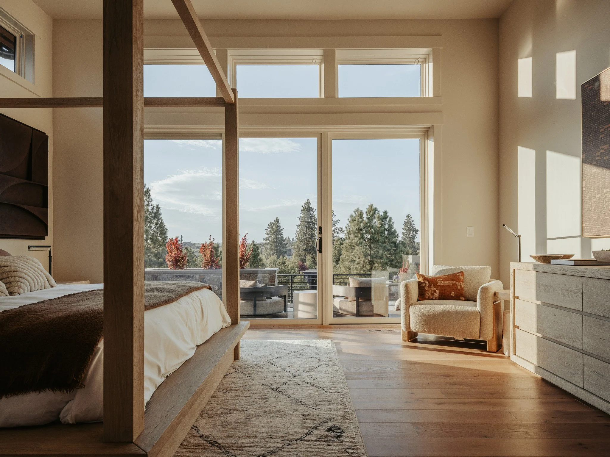 Living room with large windows showing trees outside, beige sofa with orange pillows, wooden dresser, and a view of the sky.