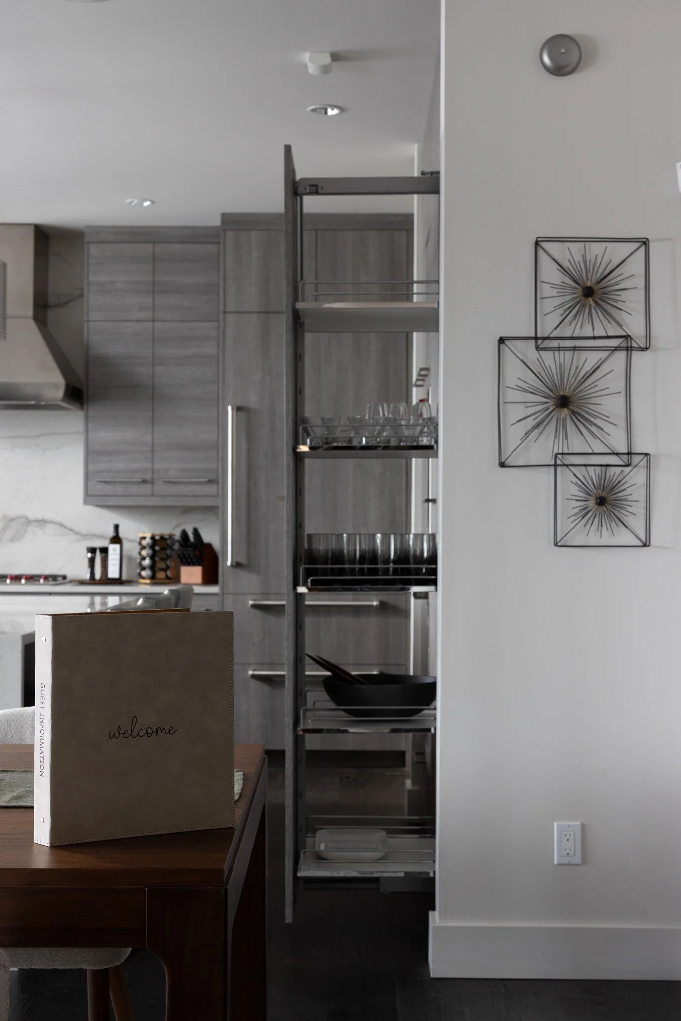 Modern kitchen with gray cabinets, a metal shelving unit, decorative wall art, a welcome sign, wine bottles, and glassware.