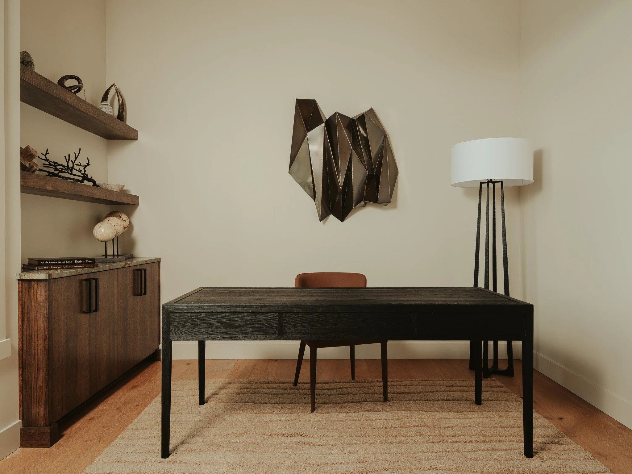 Interior room with a dark wooden table, black chair, a white standing lamp, wooden shelves with decorative items, and a geometric wall art piece.