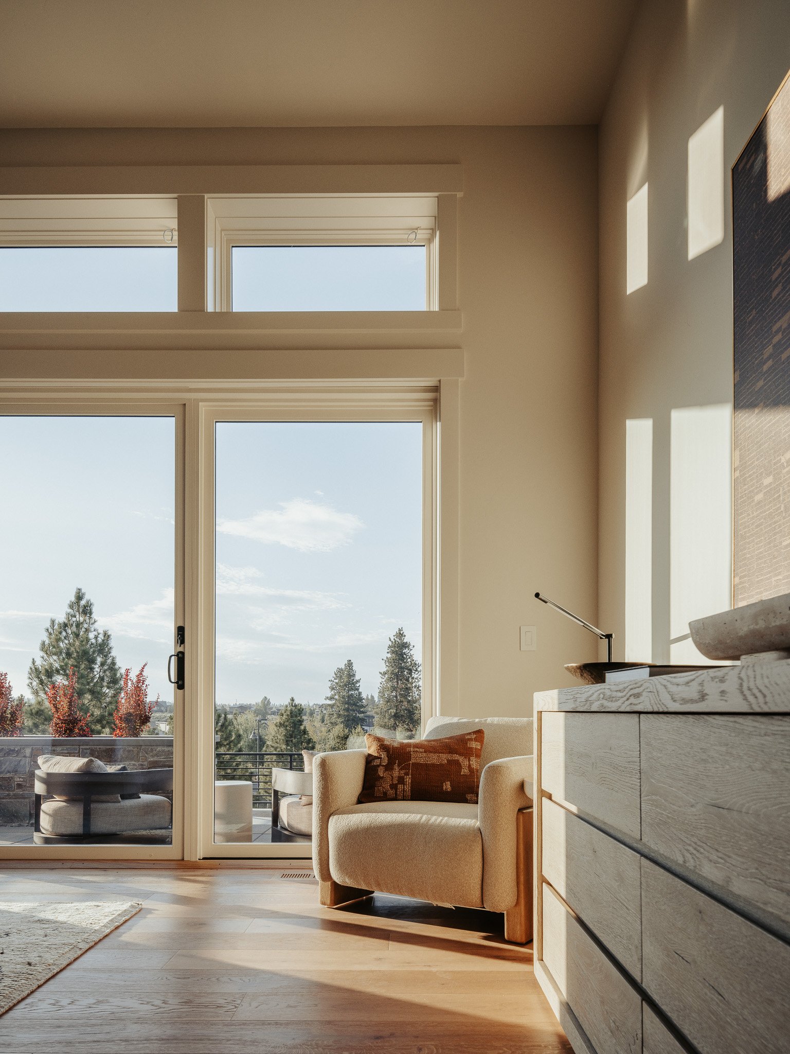 Living room with large glass sliding door, beige sofa with orange pillow, wooden dresser, and scenic outdoor view of trees and sky.