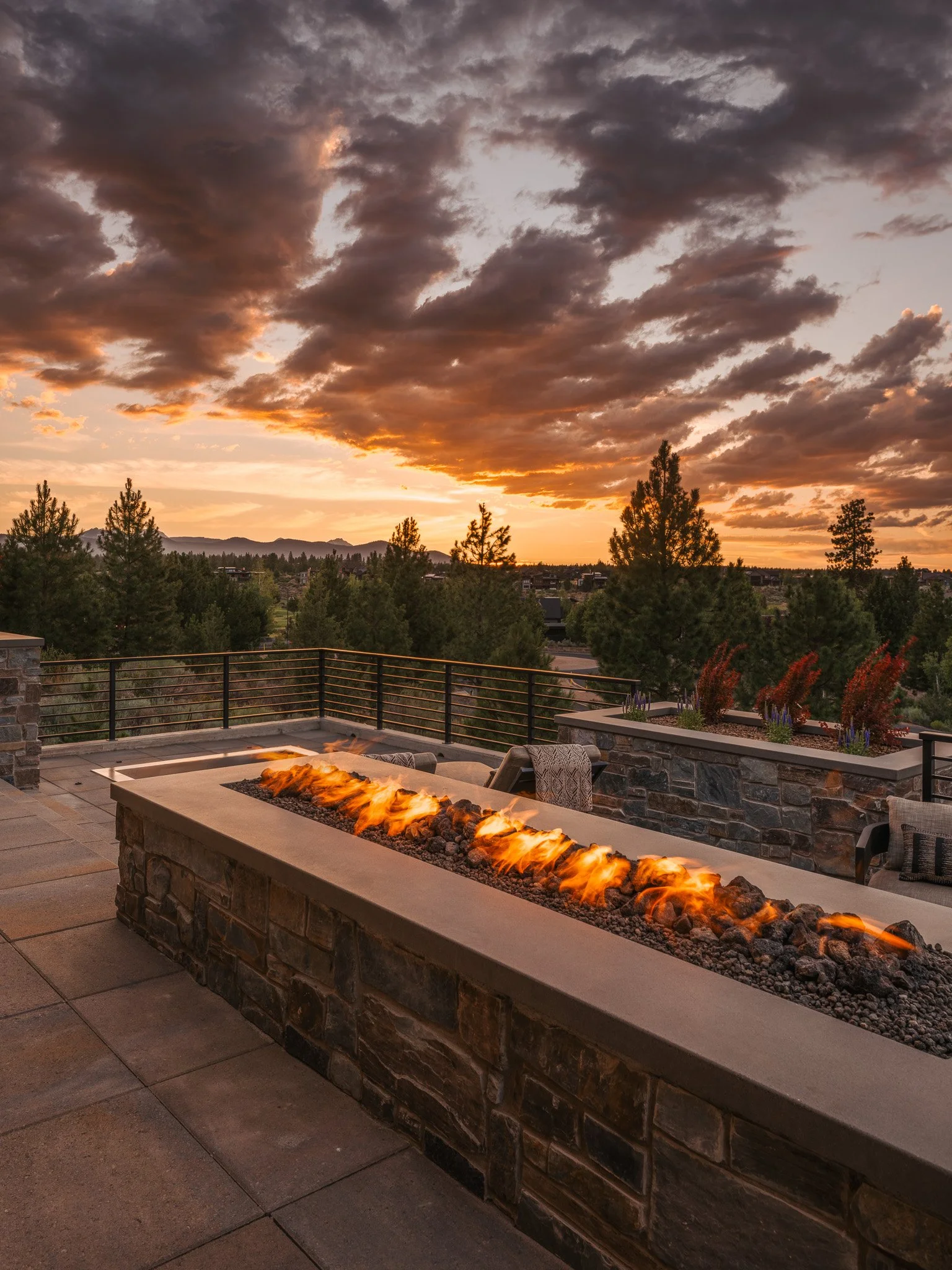 Outdoor patio with a modern fire pit, surrounded by chairs, overlooking a scenic sunset with cloudy sky and distant mountains.