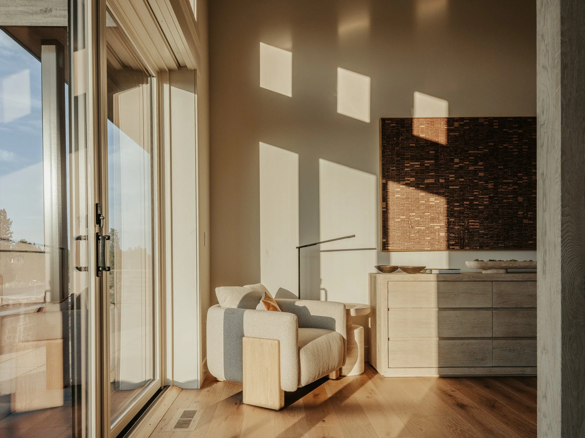 Living room with a beige sofa, a wooden coffee table, and a wall-mounted television. Large glass sliding doors with sunlight streaming in, casting rectangular shadows on the wall. Wooden flooring and a textured brick wall background.