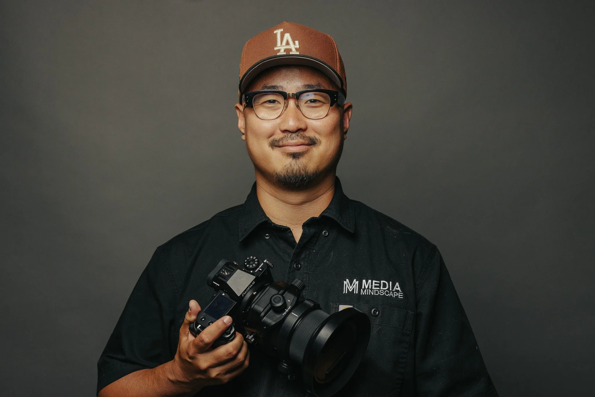 A man wearing glasses and a brown LA cap holding a professional camera, standing in front of a plain gray background.
