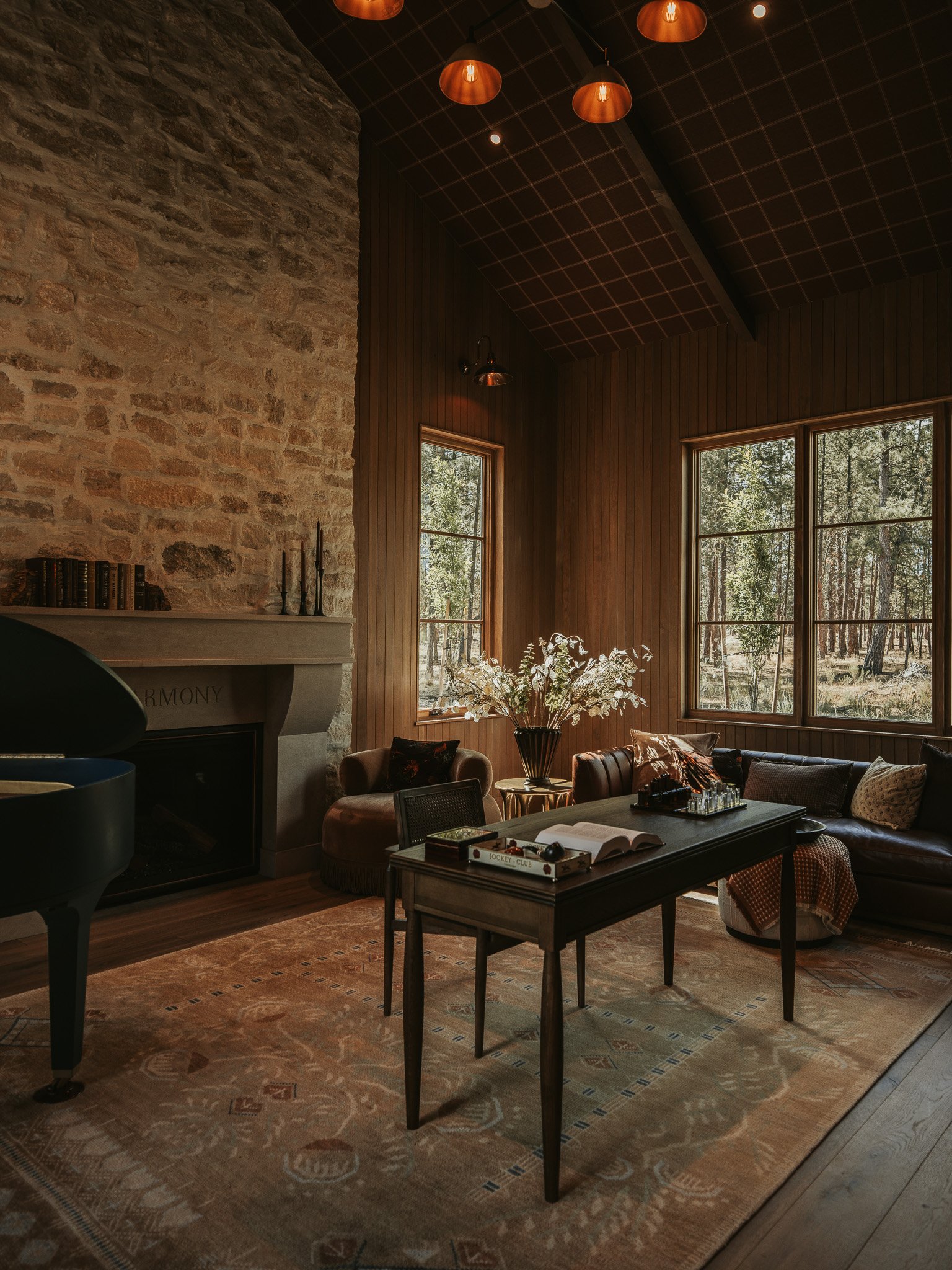 Living room with stone and wood walls, large windows showing trees outside, a dark wood coffee table with books and decorative items, a beige rug, a leather sofa with colorful pillows, a plant in a black vase, and pendant lights hanging from the ceil
