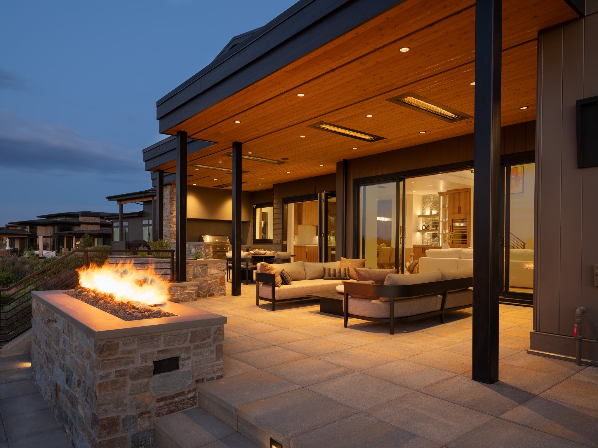 Modern house exterior with covered patio, outdoor fireplace, seating area, and string lights at dusk.
