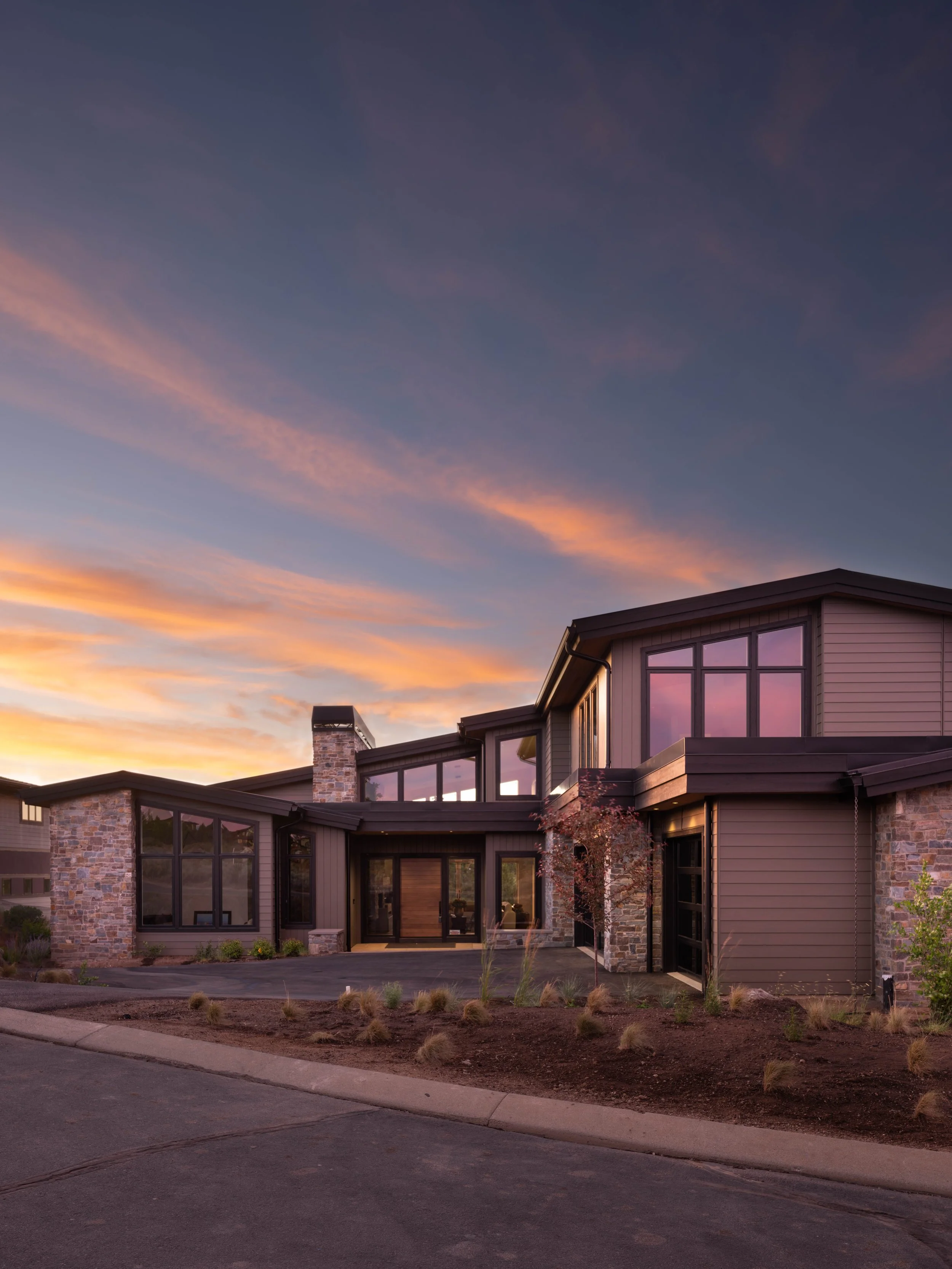 A modern house with stone and siding exterior, large windows, and a mix of flat and sloped rooflines, set against a colorful sunset sky.