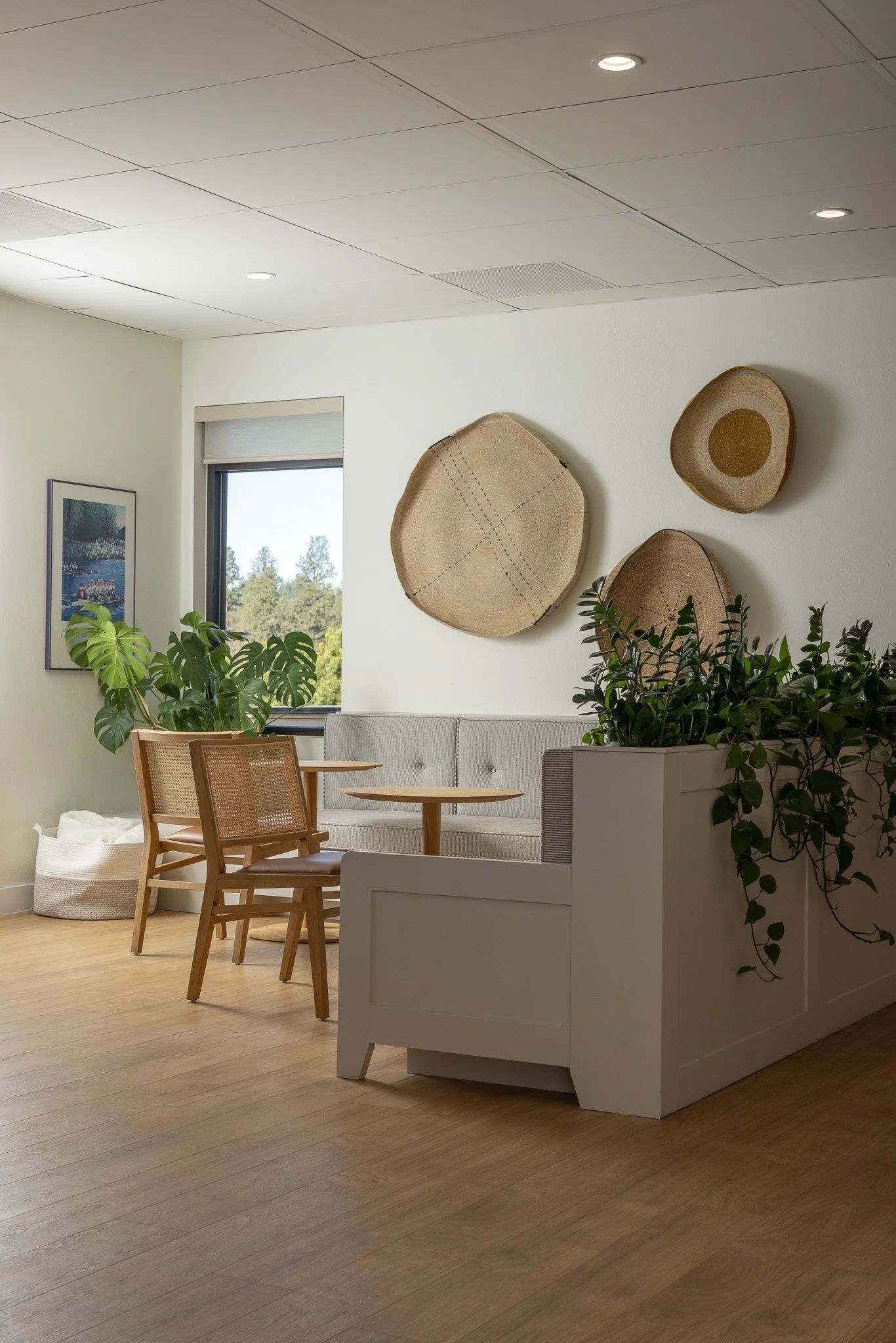 Interior of a bright room with a window showing trees outside. The room has a white wall decorated with three woven wall hangings, a framed picture, and green plants. There is a white sofa, wooden chairs, a small round wooden table, and a white stora