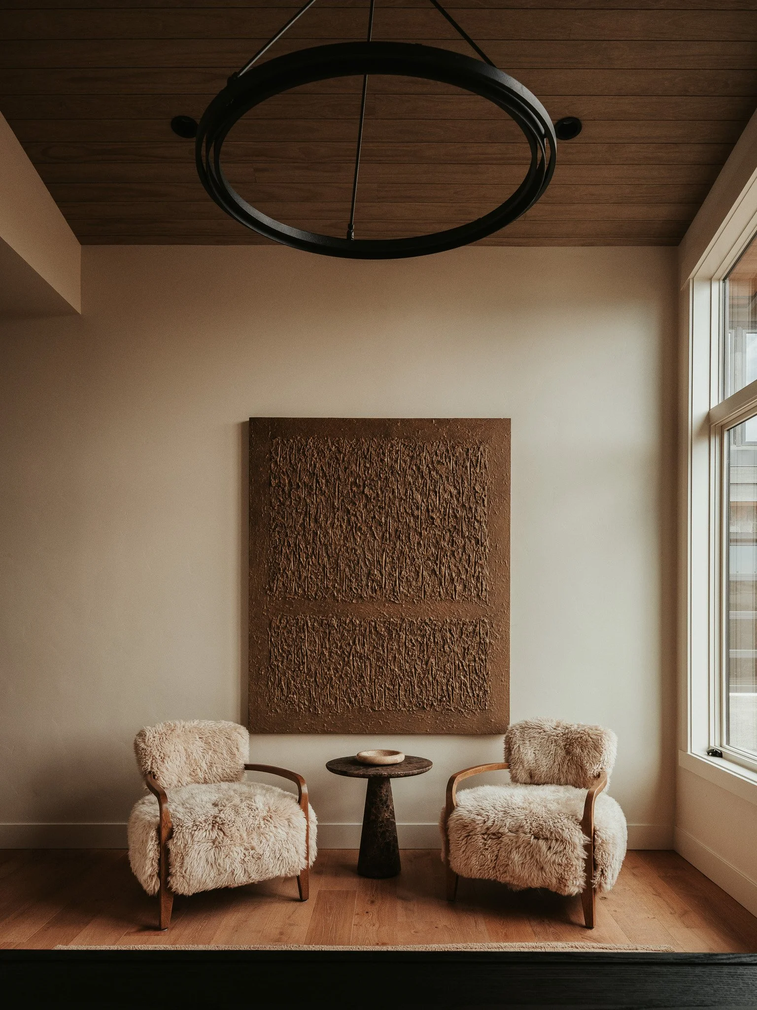 Two armchairs with cream furry upholstery facing each other with a small black round table between them, a textured brown artwork on the beige wall behind, and a window on the right side of the room.