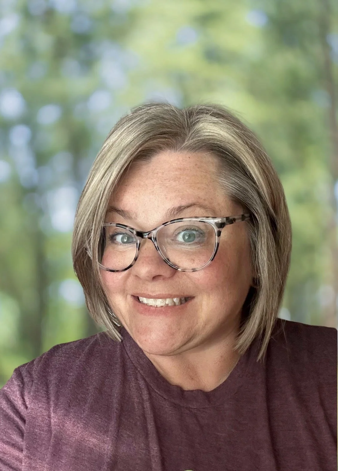 A woman with glasses and shoulder-length hair smiling, outdoors with a blurred leafy background.