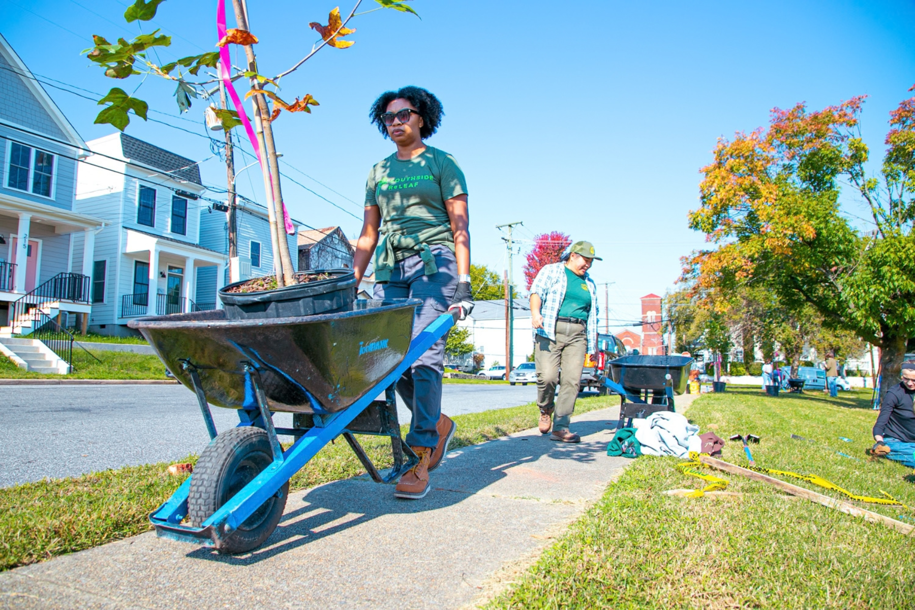 Sheri transports a potted tree in a wheelbarrow to its planting site and Qui gathers supplies in the background.