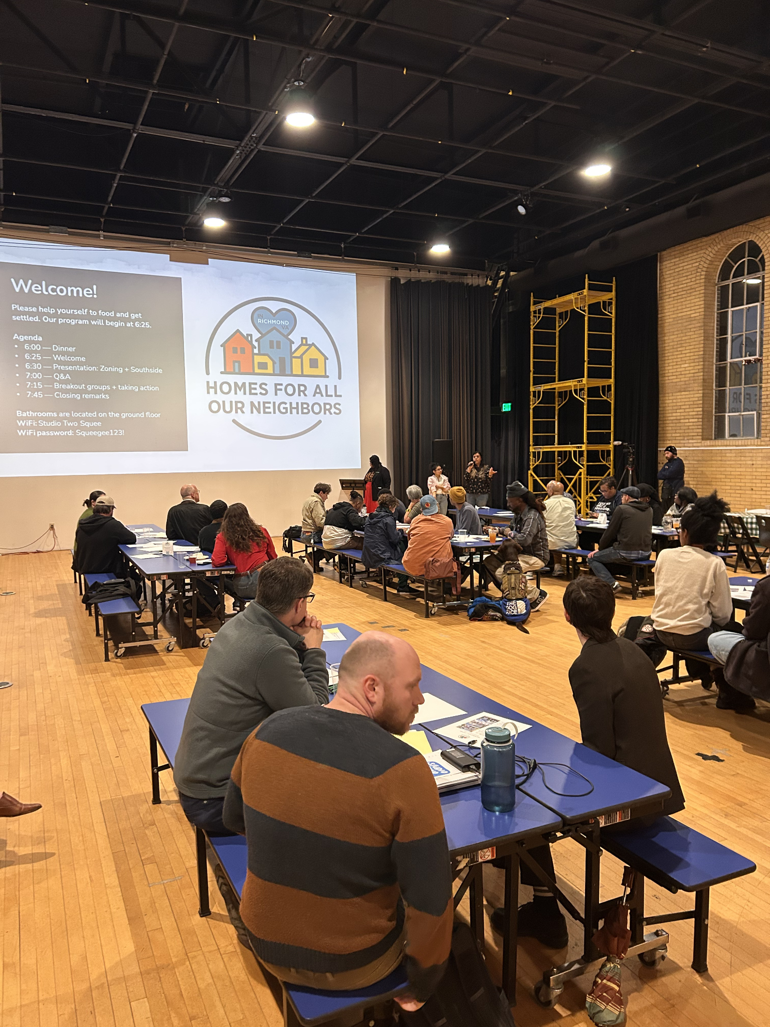 A group of people sitting at tables in a large room attending a community event or meeting. There is a large screen displaying a presentation about a program called "Homes for All Our Neighbors" with a logo featuring three houses and a heart. The room has wooden floors, brick walls, and high windows, with some individuals speaking at a podium and others listening.