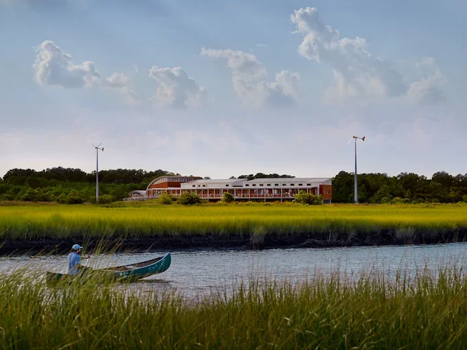 Photo of the Brock Environmental Center from across the Lynnhaven River, where a person in a ball cap rows a canoe on a sunny day.