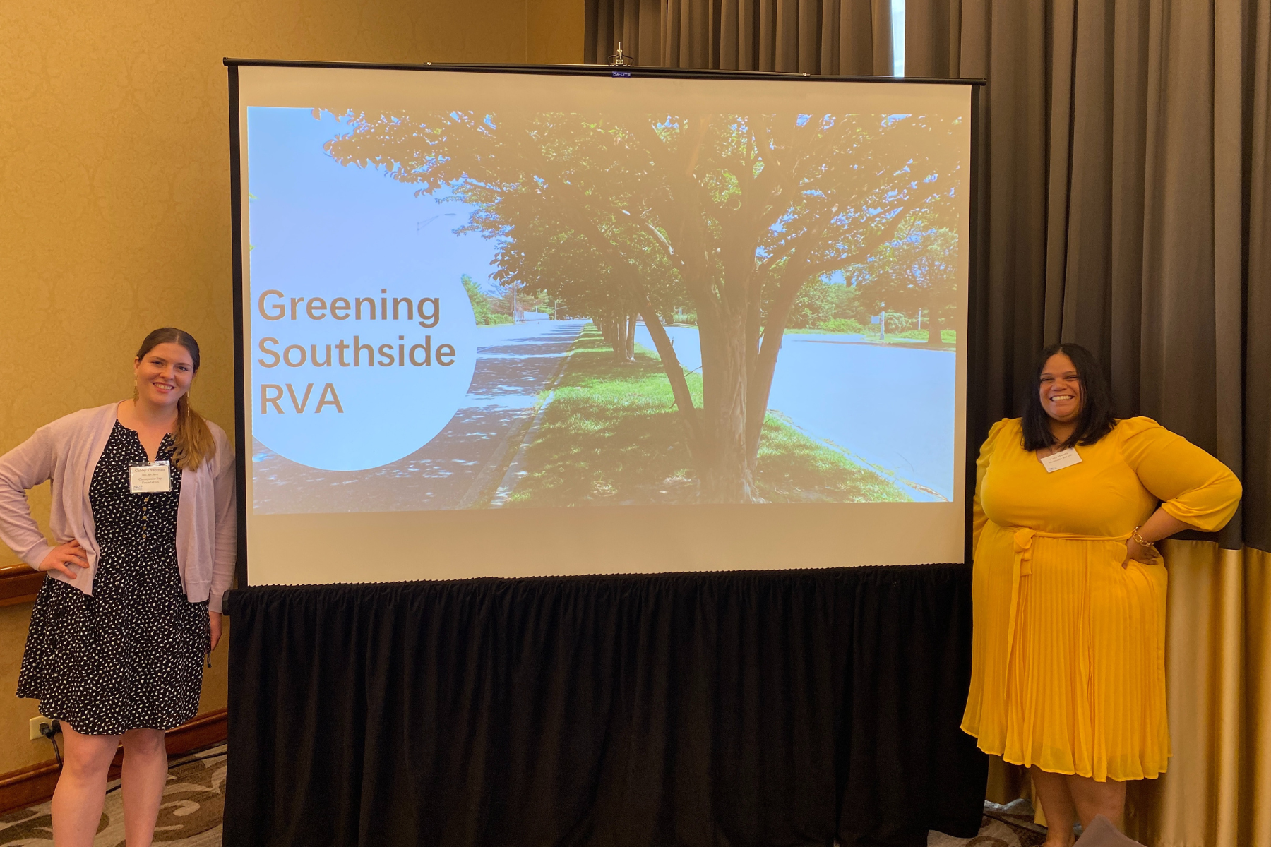 Two women standing on either side of a presentation screen that displays a park scene with trees and a sidewalk, with the text 'Greening Southside RVA'.