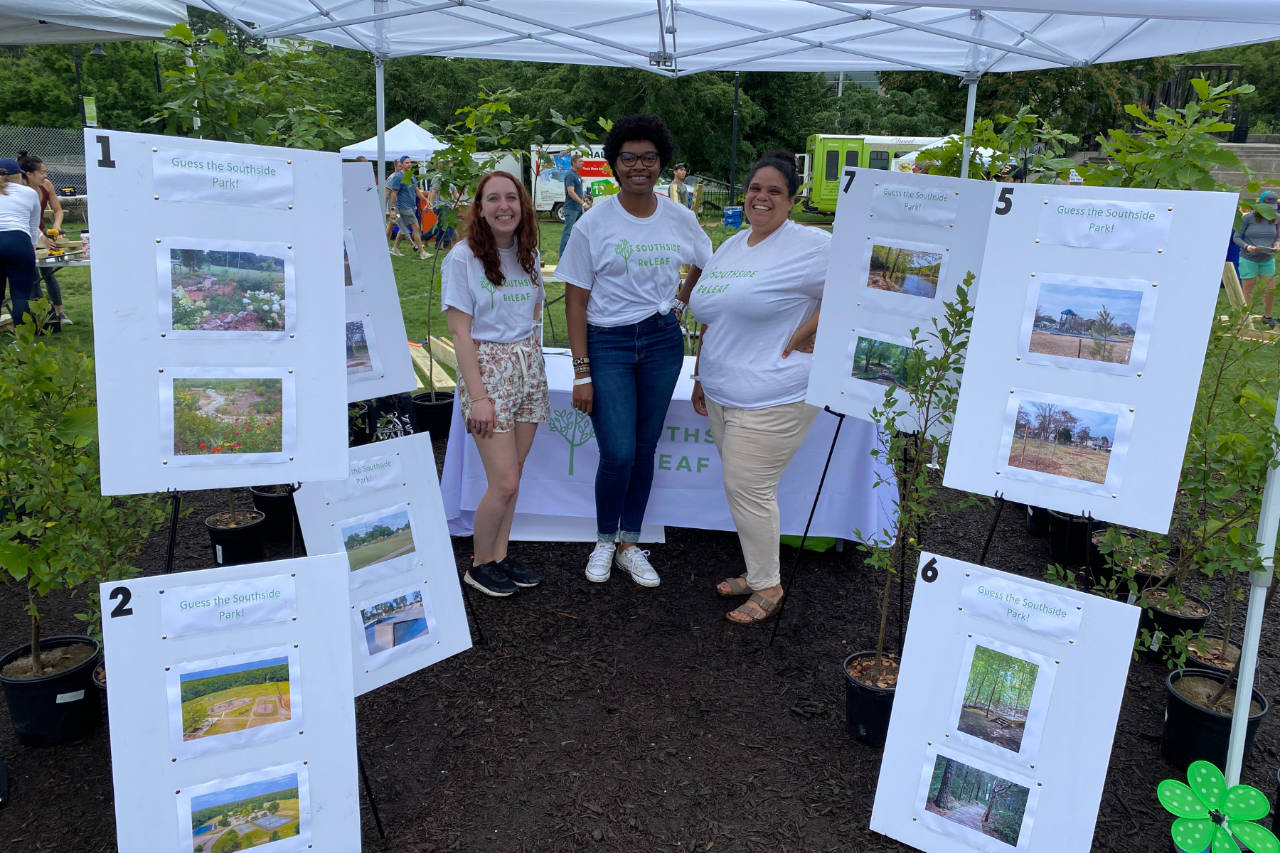 Three women in white shirts standing in front of display boards with pictures of parks, at an outdoor community event. Event tents and other people can be seen in the background.