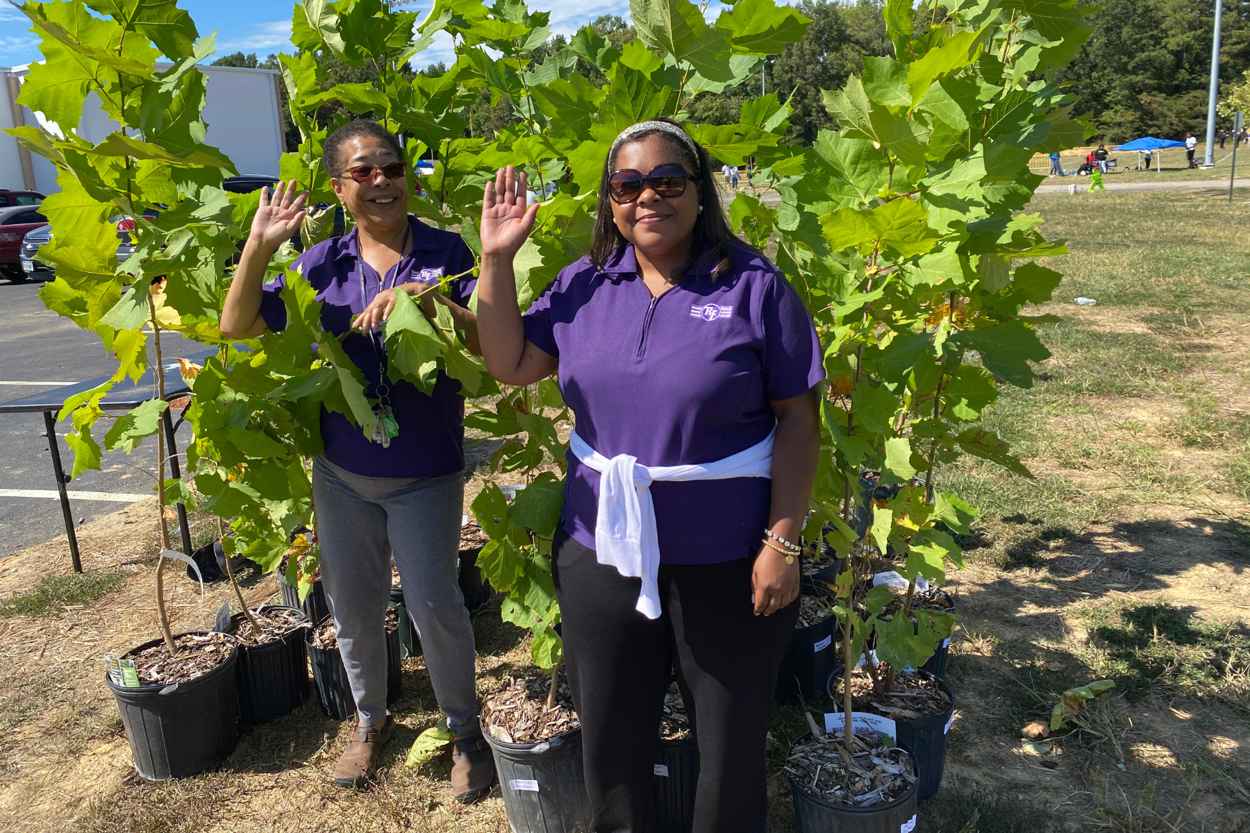 Two women in matching purple shirts wave to the camera while posing in front of a stand of potted trees at our tree giveaway.