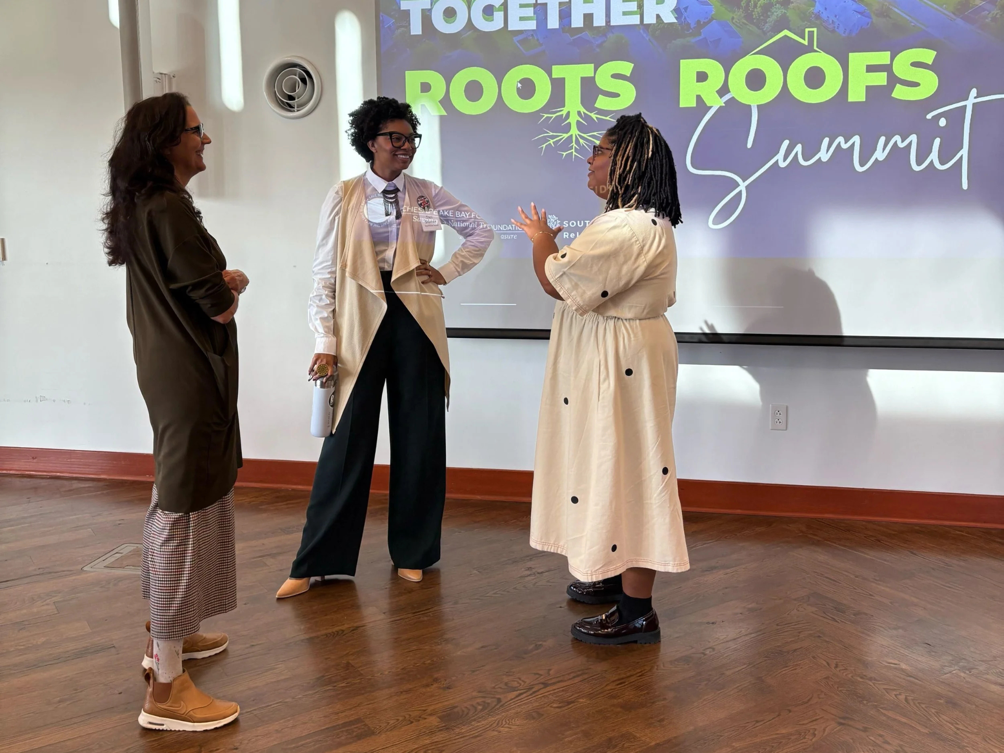 Lisa Renee Jennings, Sheri Shannon, and Kendra Norrell talking in front of a projector screen showing the Growing Together Roots to Roofs graphic at the Brock Environmental Center.