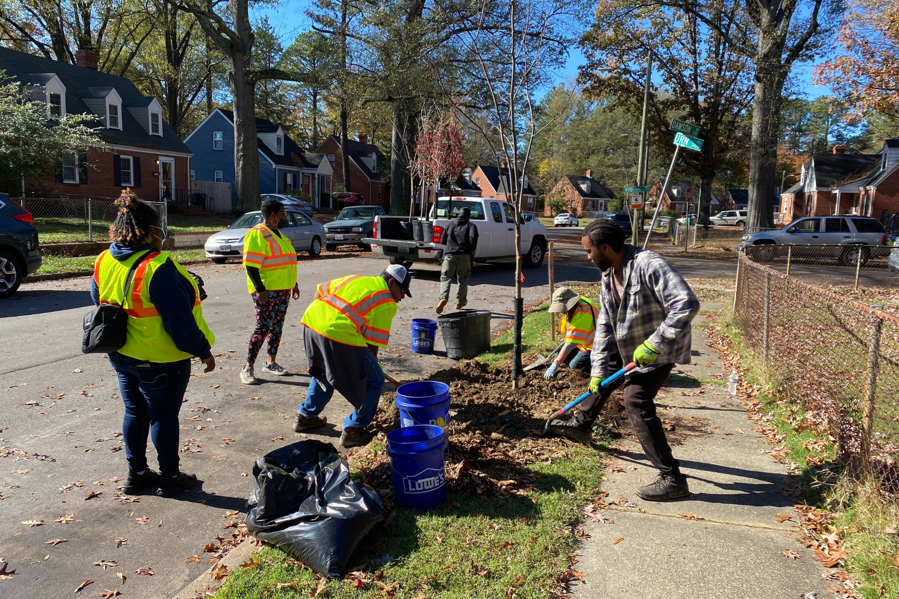 Group of people planting trees along a residential sidewalk on a sunny day, some wearing yellow safety vests, others with gardening tools, and a truck parked on the street.