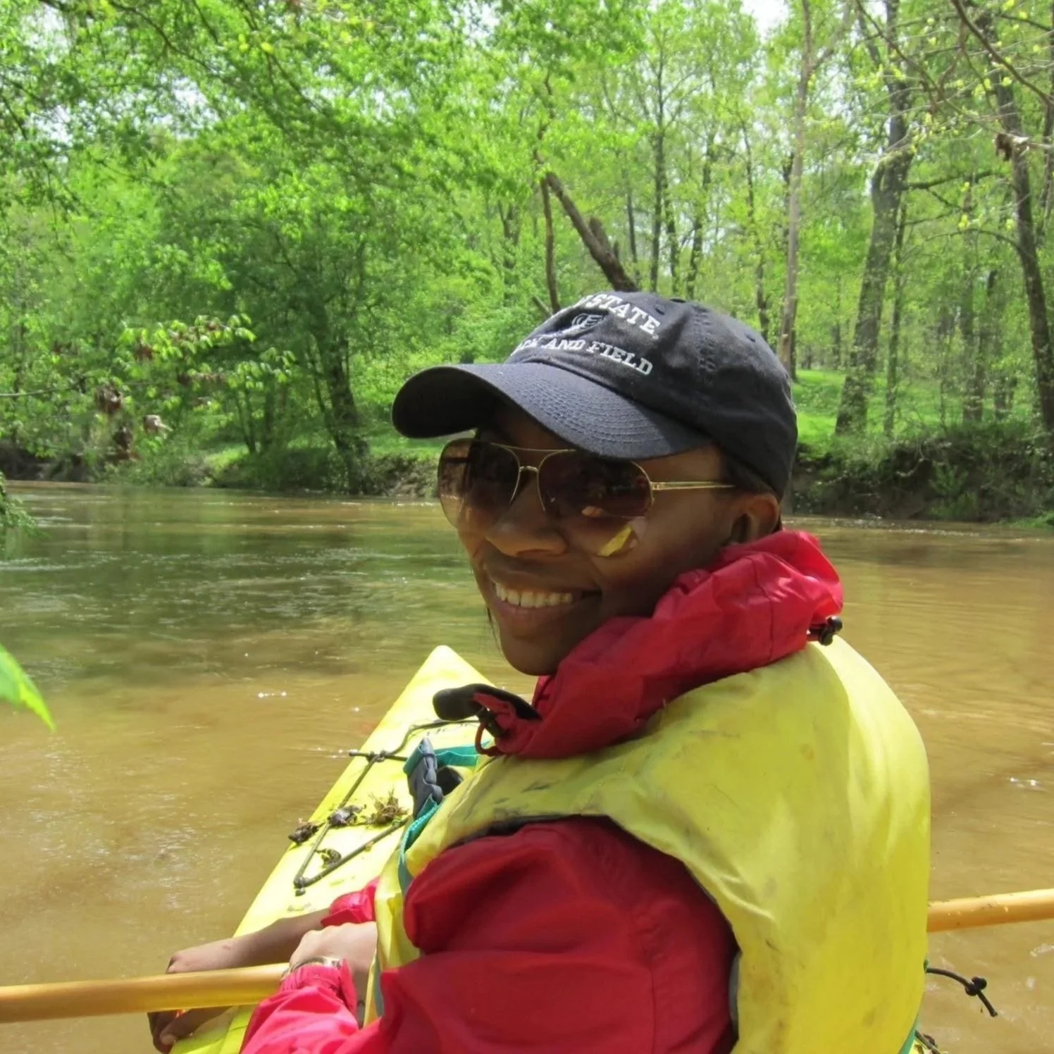 Sheri smiles while sitting in a kayak on a muddy river, wearing a baseball cap, sunglasses, and a life jacket, with a dense forest lining the riverbank in the background.