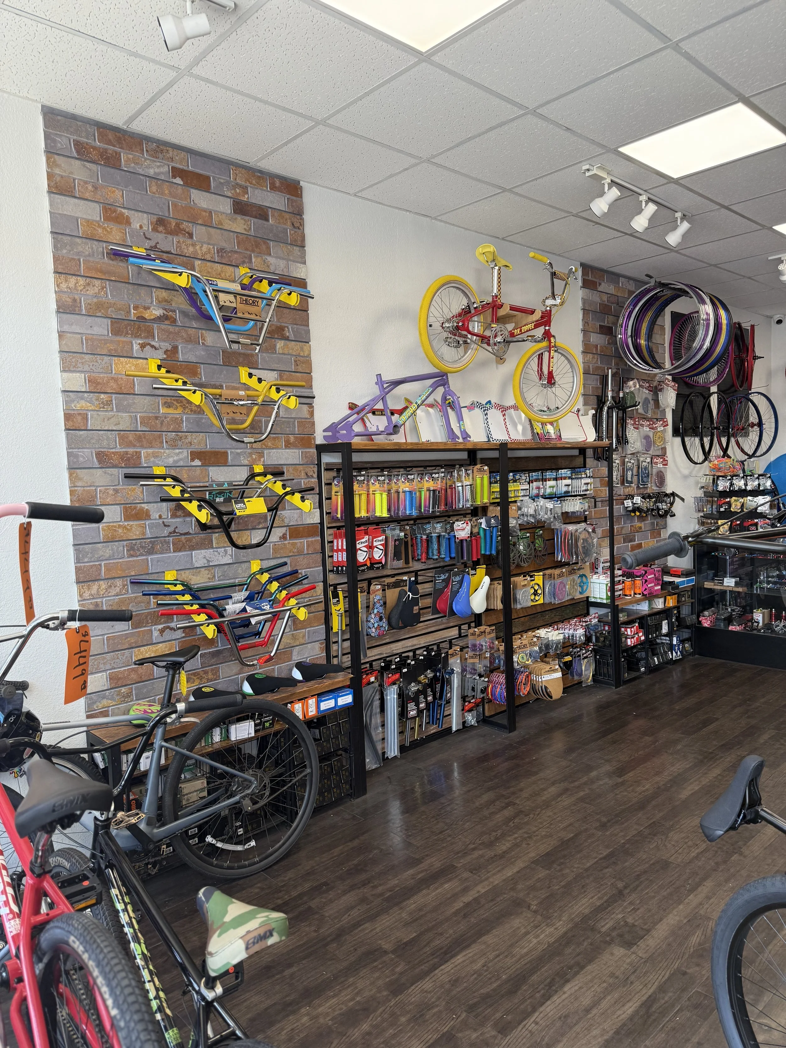 A bicycle shop with various bikes, bike parts, and accessories on display. There are wall-mounted bikes, hoops, and bike frames, along with shelves stocked with tools, helmets, water bottles, and other cycling gear. BMX handlebars. Wheelie. Bikelife.
