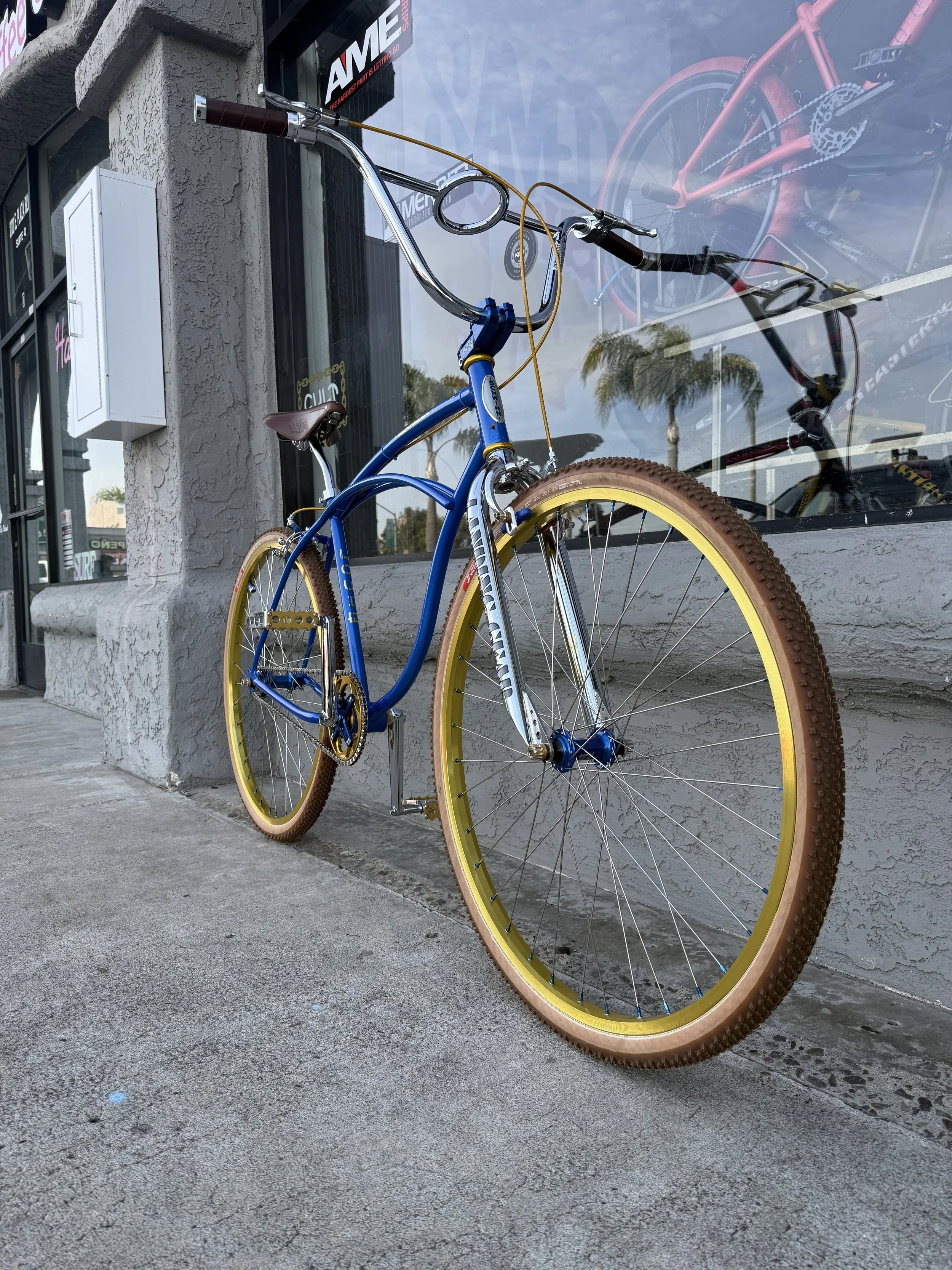 A vintage-style bicycle with a blue frame, yellow tires, and brown saddle, leaning against a gray building with a large window displaying a pink and red bicycle.