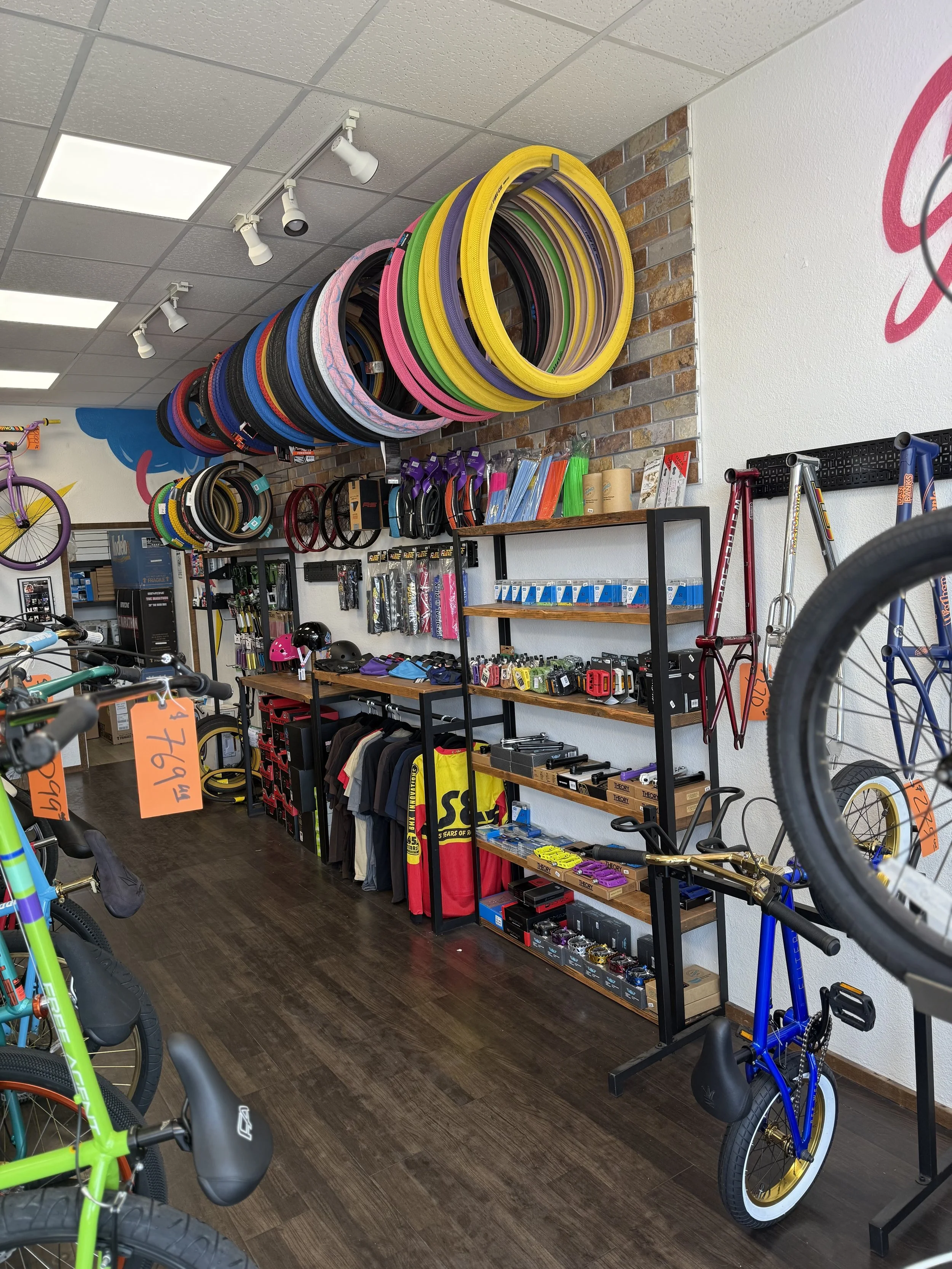 Indoor bicycle shop displaying various bicycle accessories, helmets, and bikes, with colorful bike tires arranged on the wall and shelves with bike gear and parts. Wheelie. Bikelife. BMX. SE. Throne. Mafia. GT.