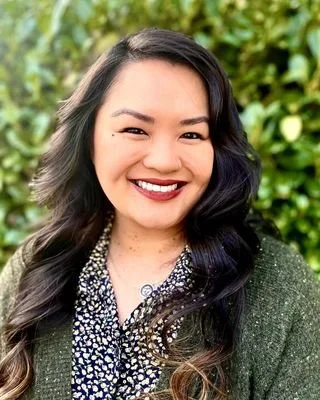A woman with long dark hair smiling outdoors, wearing a patterned blouse and a dark green jacket, with a background of green foliage.