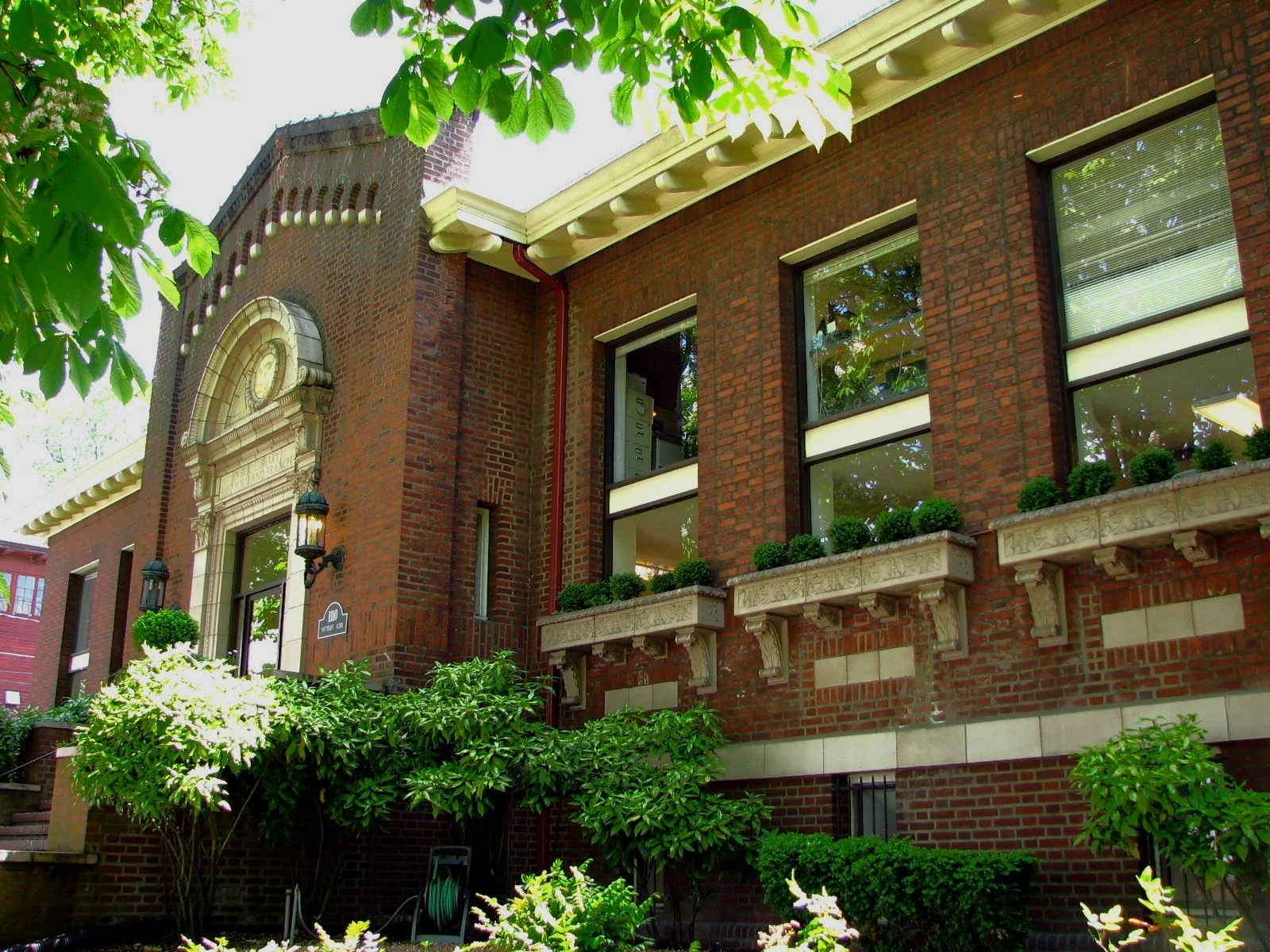 A red brick building with large windows and decorative stone planters with green bushes, surrounded by lush green trees and plants.
