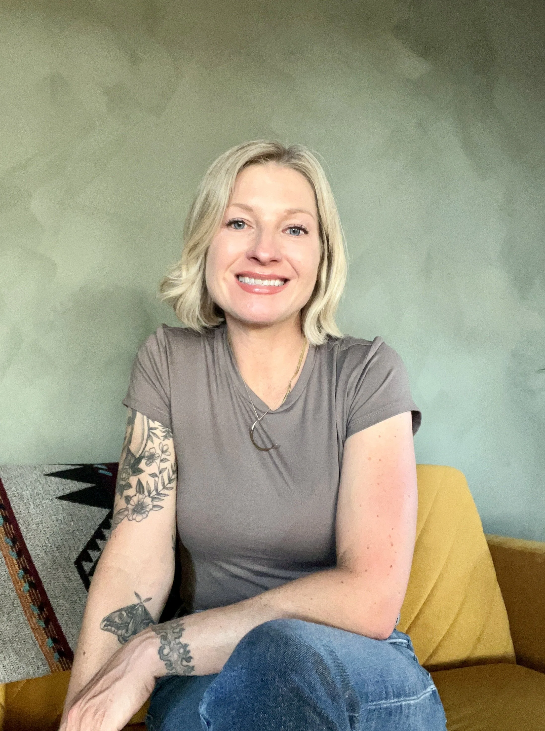 A woman with blonde hair smiling, wearing a gray t-shirt, sitting on a yellow couch with a green wall in the background.