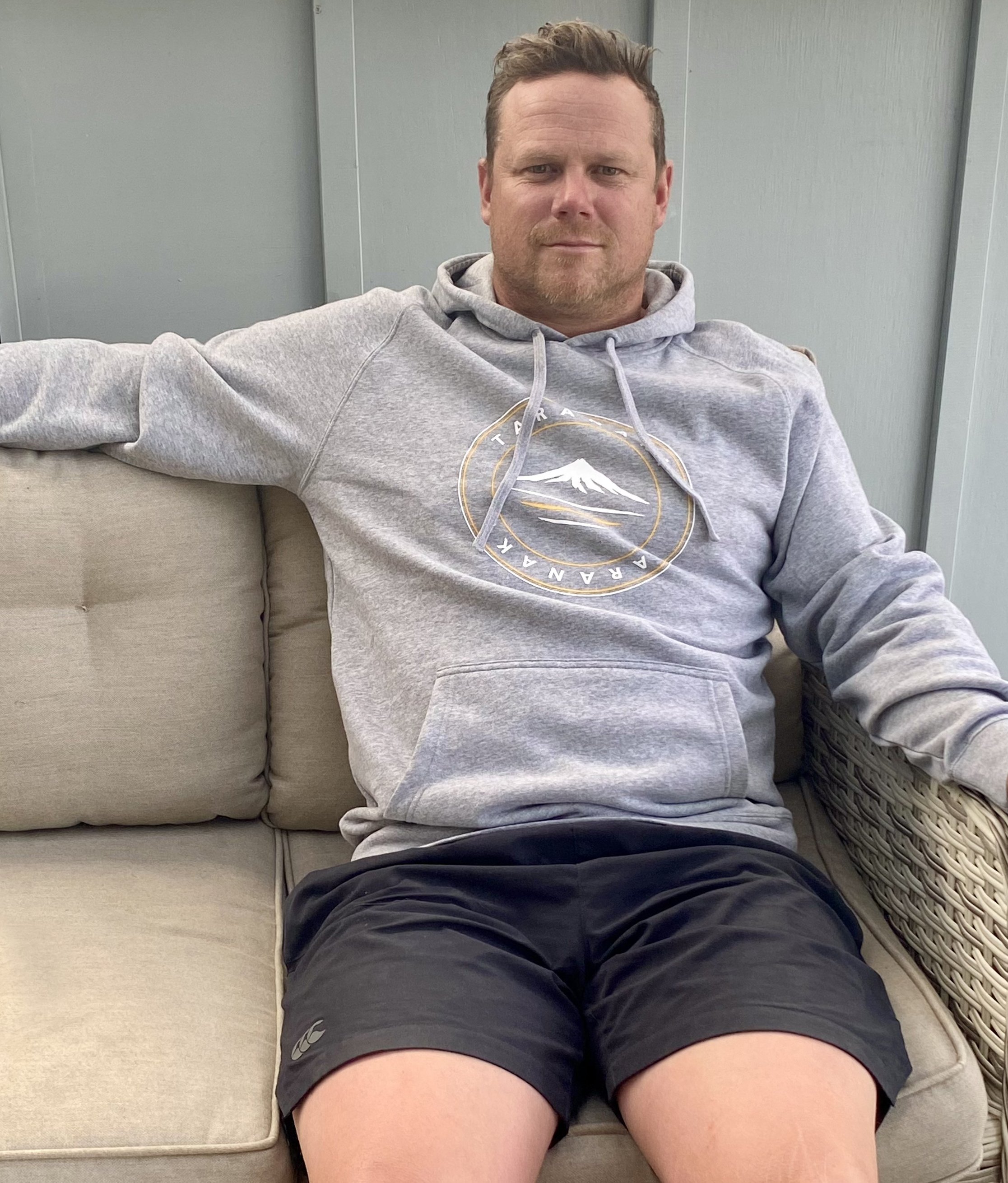 Man relaxing in chair wearing the grey marle Adults Taranaki Classic Logo Hoody.