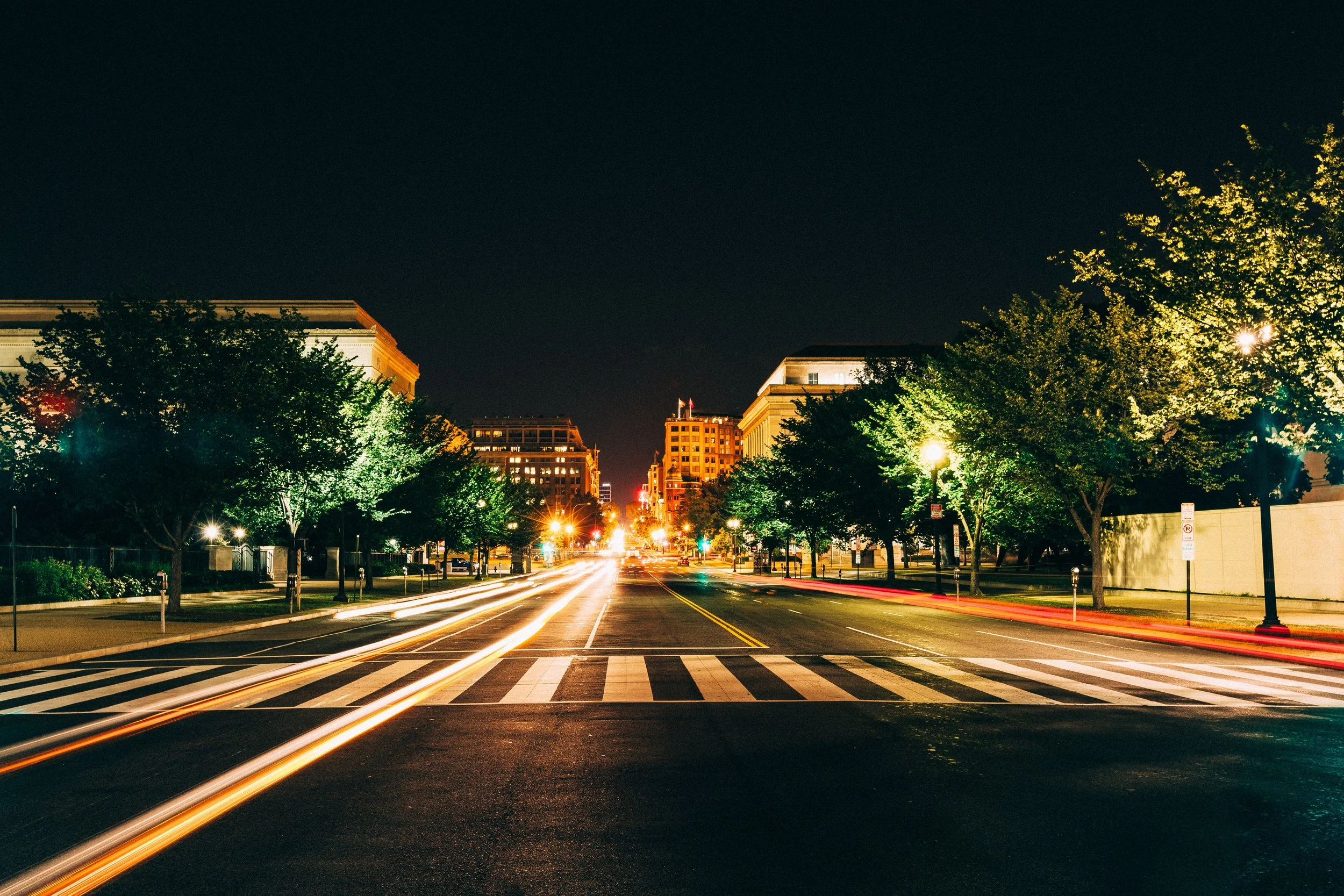Symmetrical city avenue at night with crosswalks, trees, streetlights, and white and amber traffic trails